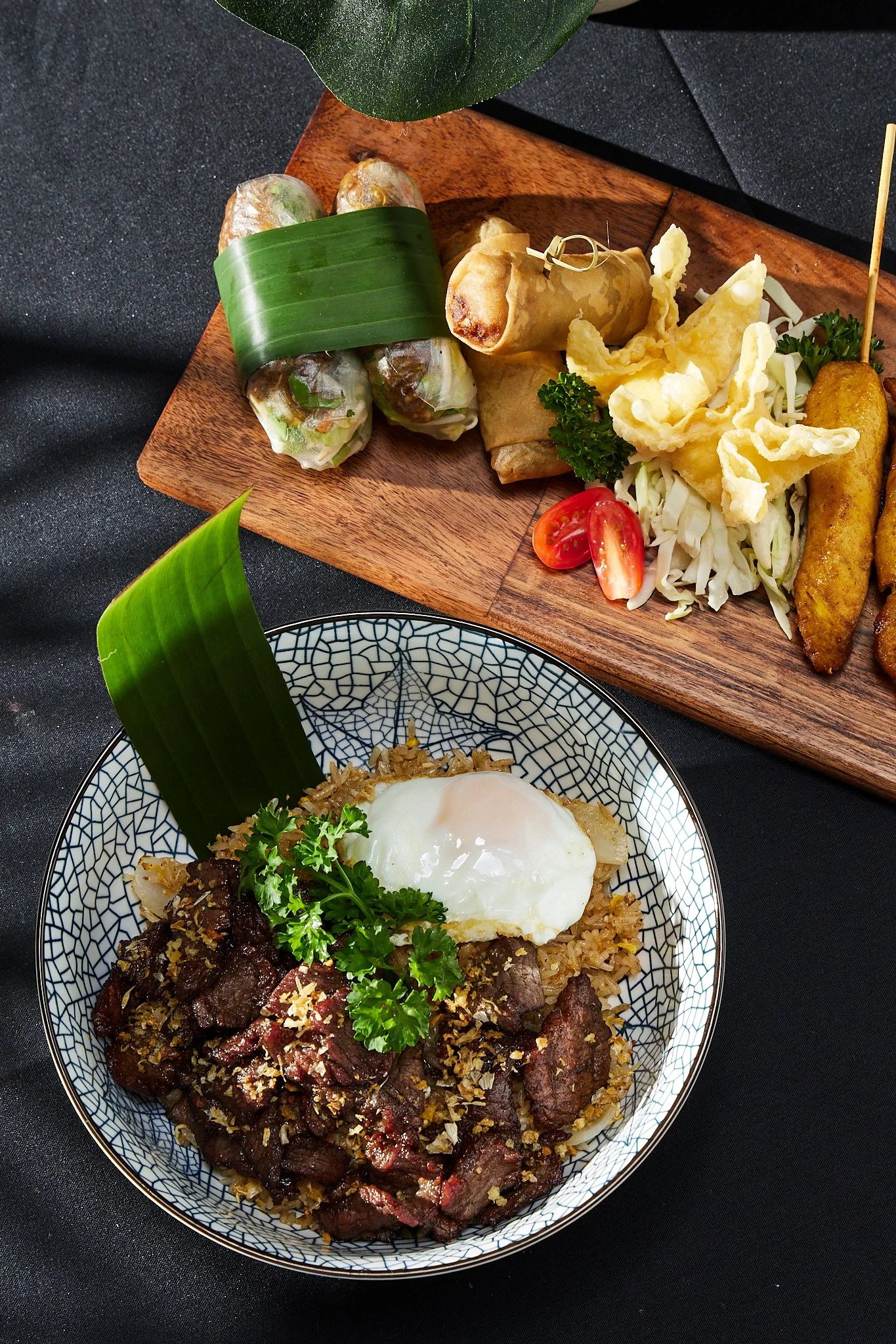 A bowl of beef stew topped with a poached egg and garnished with parsley, with a piece of banana leaf on the side. Above, a wooden platter with spring rolls, fried snacks, and vegetables.