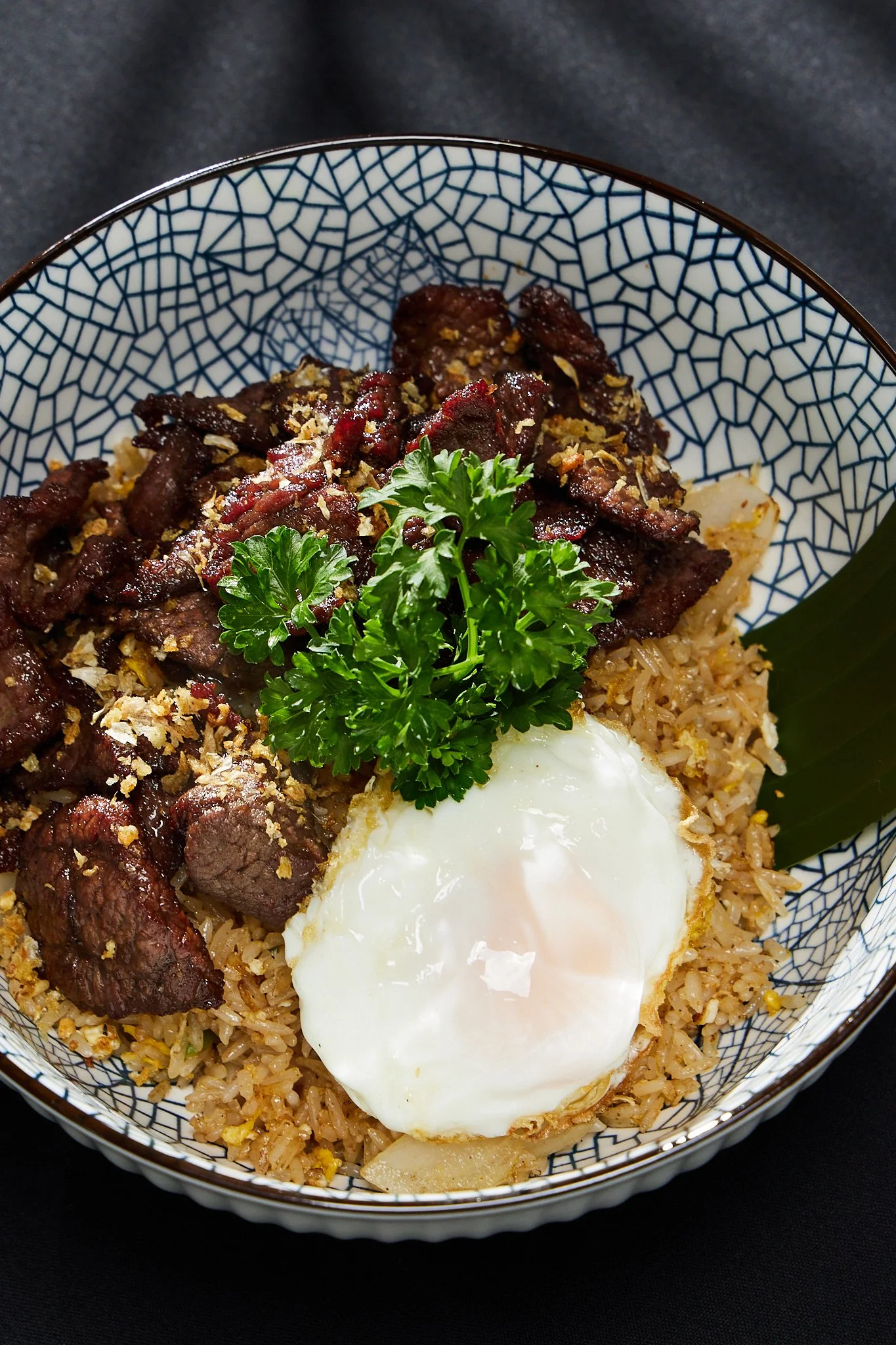 A bowl of fried rice topped with cooked beef strips, a fried egg, fresh parsley garnish, and a green leaf, served in a blue and white patterned bowl on a black background.