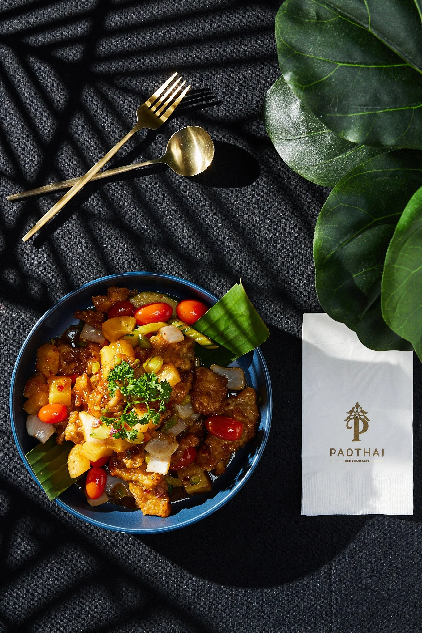 A bowl of Thai cuisine featuring vegetables and possibly fried protein garnished with cilantro, placed on a black cloth with a gold fork and spoon, green leaves, and a restaurant napkin with the logo 'Padthai Restaurant'.