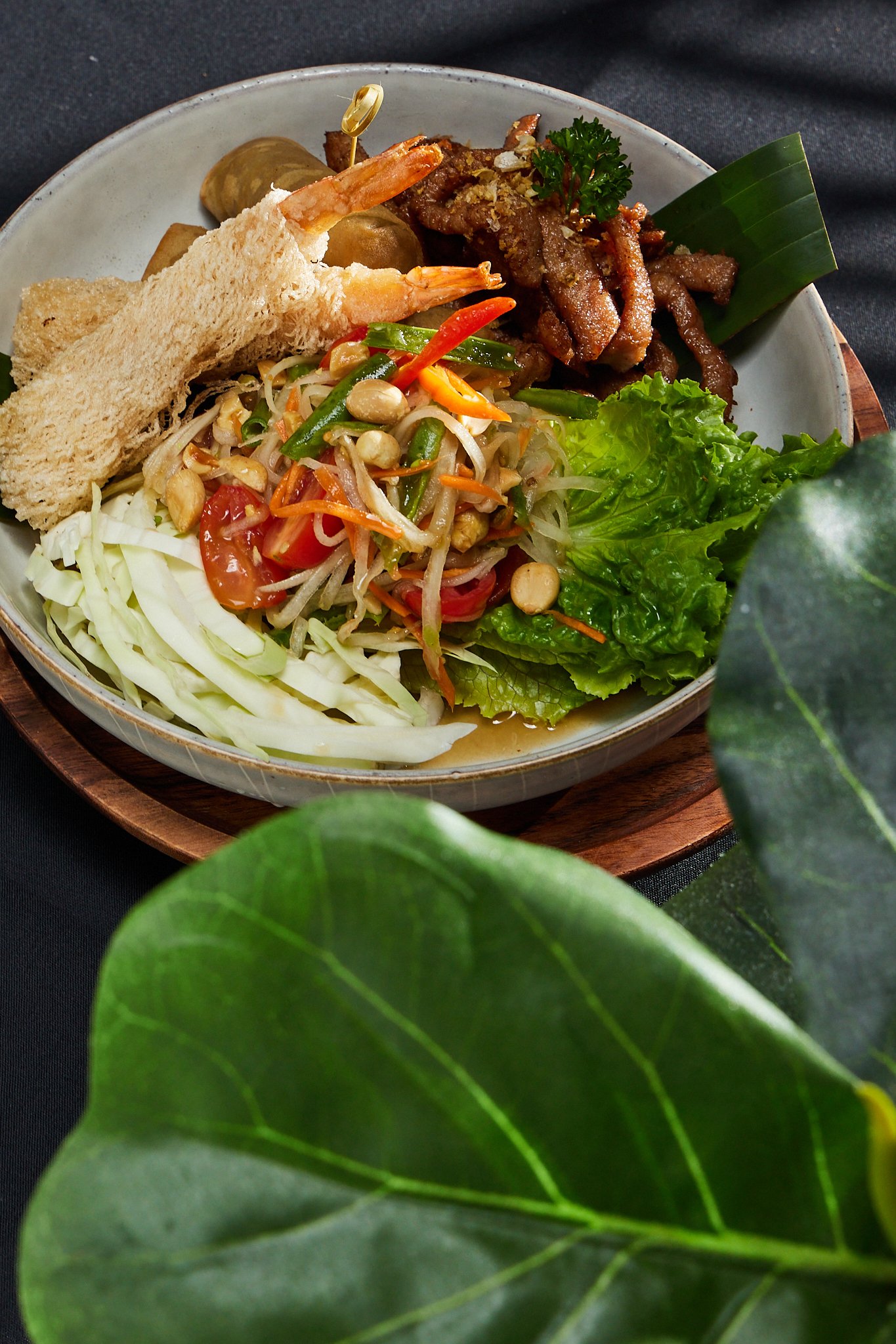 A bowl of Asian-inspired salad with shredded lettuce, tomato, bean sprouts, and chopped peanuts, garnished with cilantro, alongside fried shrimp, sliced toasted bread, and cooked beef strips, all on a wooden tray.