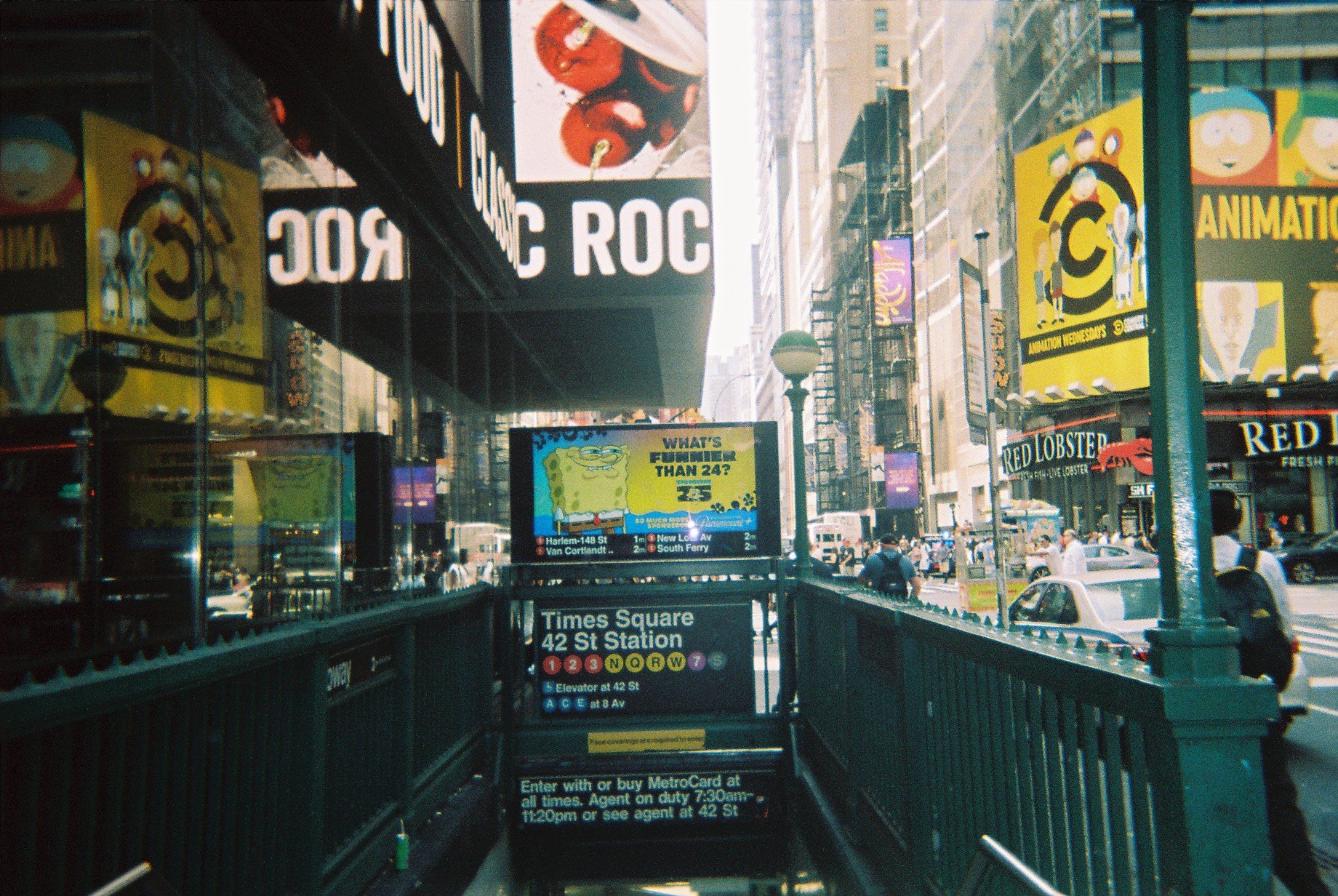 Entrance to Times Square 42nd Street subway station in New York City with stairs leading down, featuring electronic and billboard advertisements, and people walking on the street.