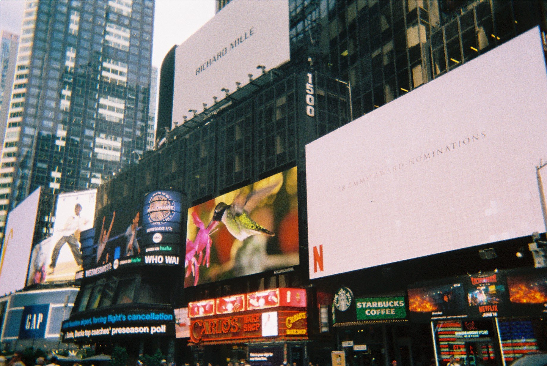 Bright digital billboards in Times Square, New York City, with advertisements for Hulu, Starbucks, and Netflix, among others. Crowds of people are visible at street level, surrounded by tall buildings.
