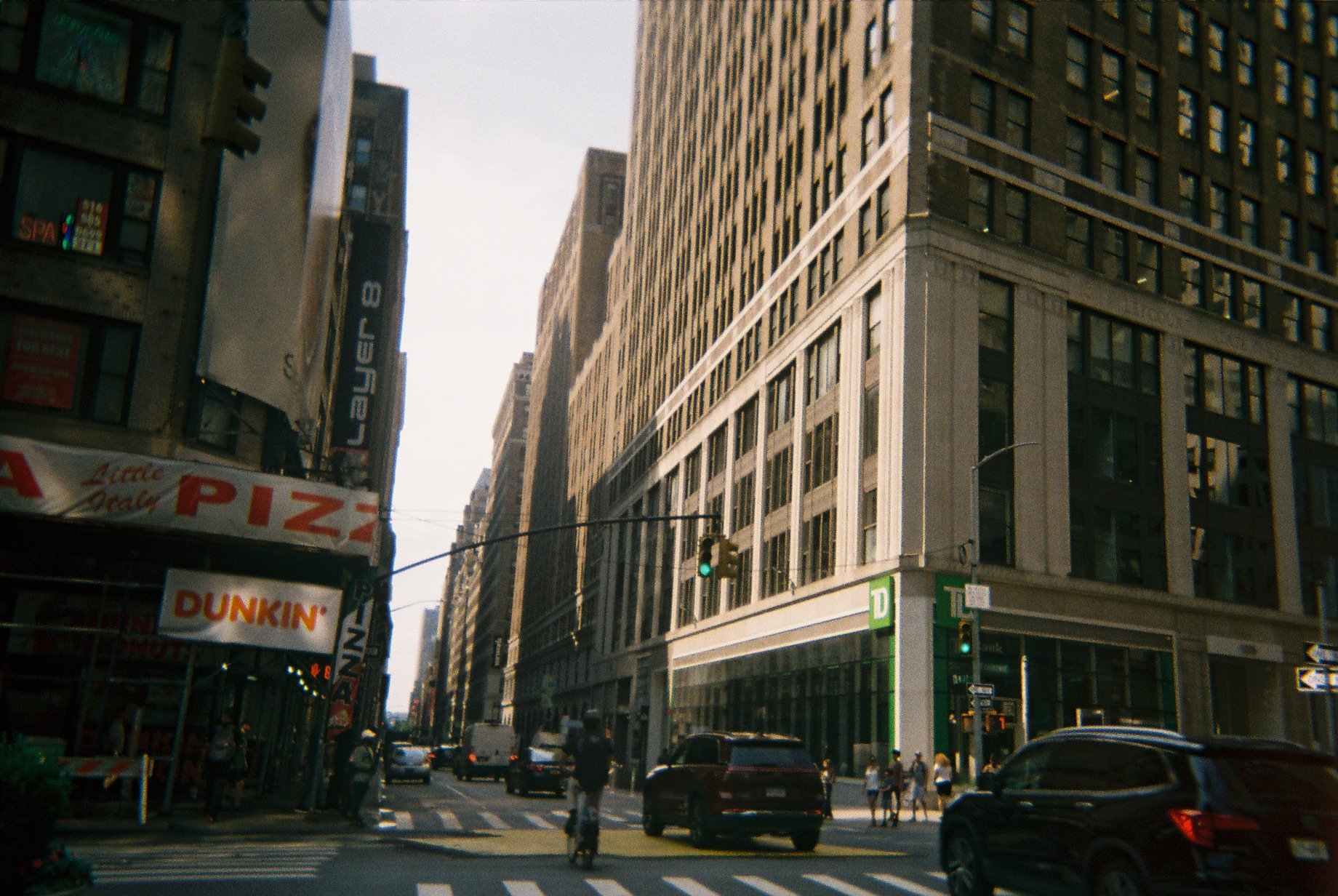 City street scene with tall buildings, cars, pedestrians, and storefronts, including a Dunkin' Donuts and a pizza shop.