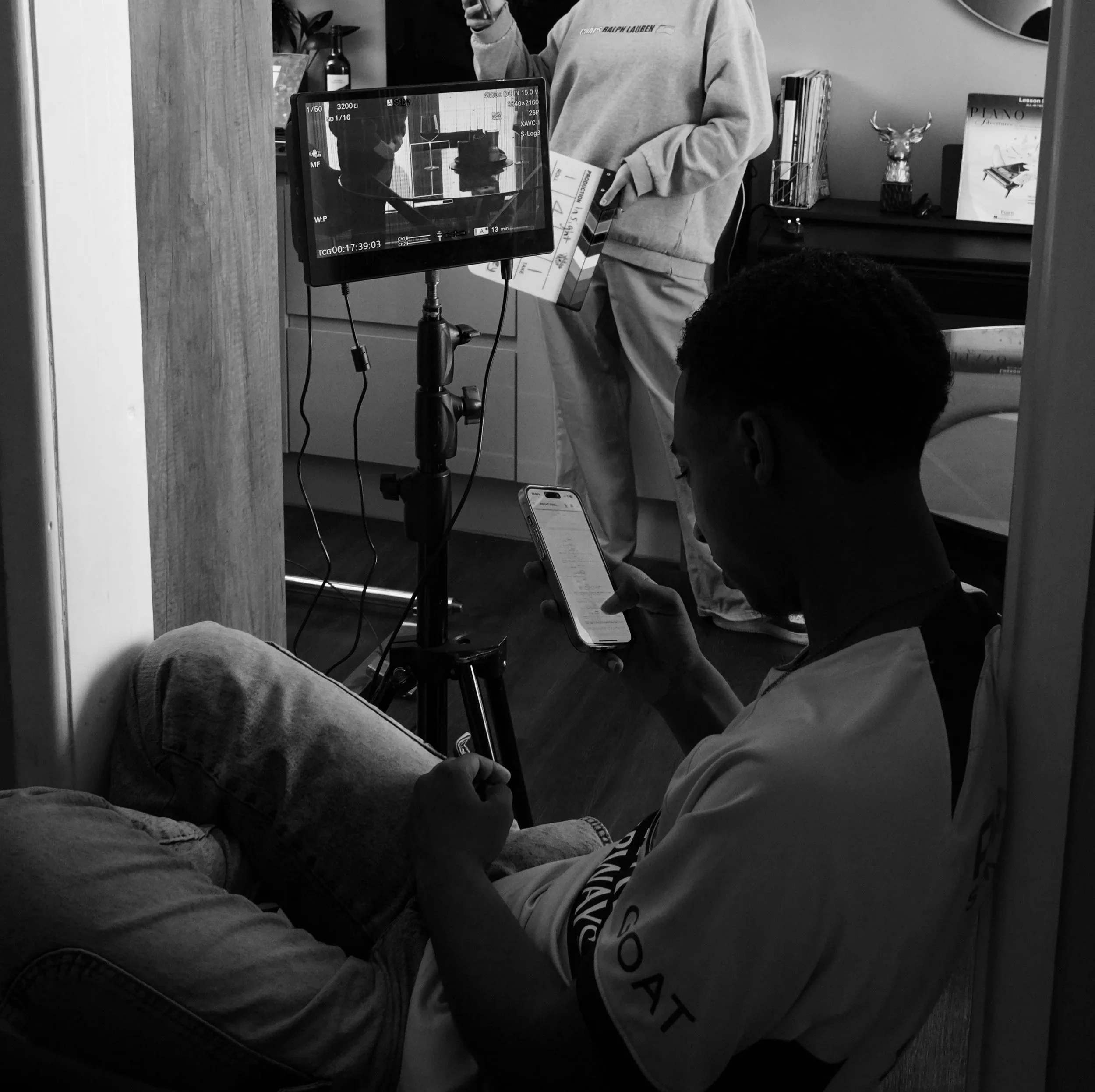 A person sitting on the floor looking at a smartphone, with a monitor camera setup in front of them capturing the scene. Another person is standing in the background near a desk with books and decorative items.
