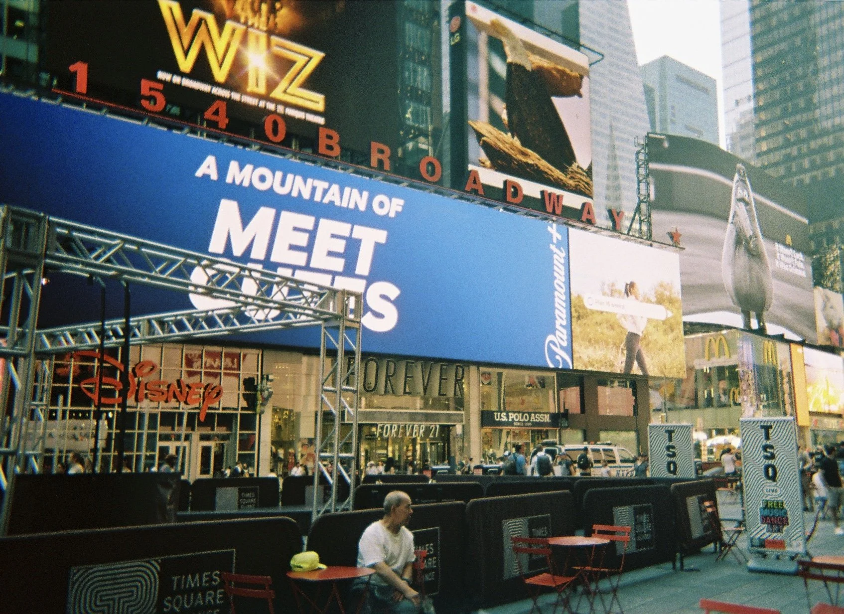 Times Square in New York City with large digital billboards and screens featuring advertisements and messages, with some people sitting at outdoor tables in the lower area.