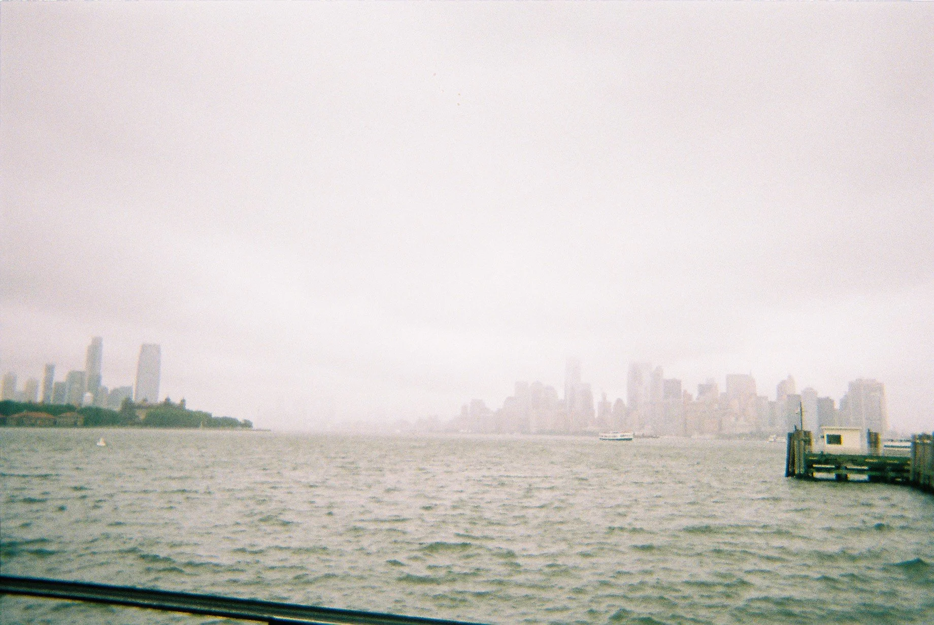 Overcast view of the New York City skyline across the water with a pier on the right and a boat in the distance.