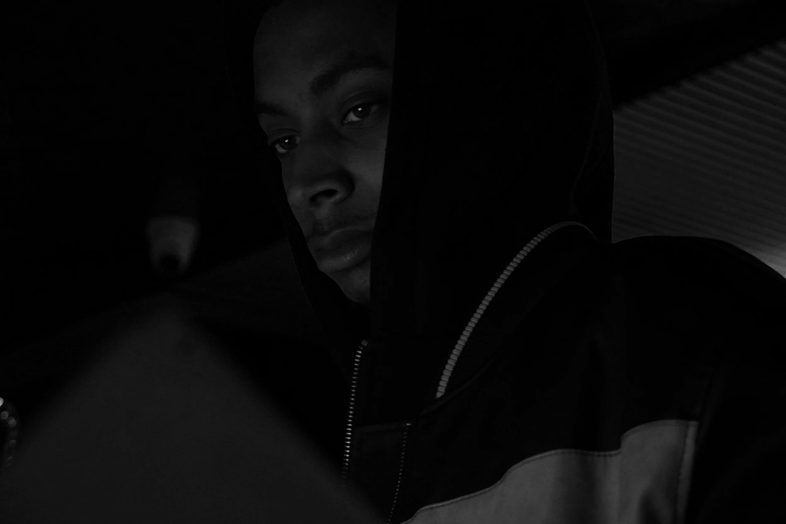 Black and white photo of a person wearing a hooded jacket, looking intently to the side, with shadows cast over part of their face.