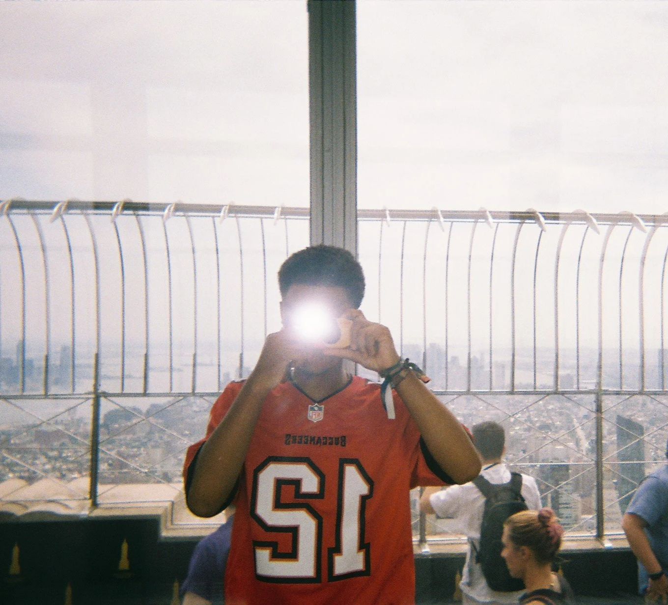Person taking a photo with a flash in front of a large window at a high vantage point, wearing an orange sports jersey with the number 12.