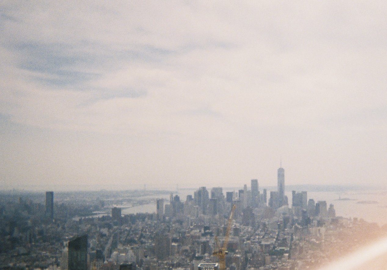 Aerial view of a city skyline with tall skyscrapers, including One World Trade Center, and a river in the background, under a cloudy sky.