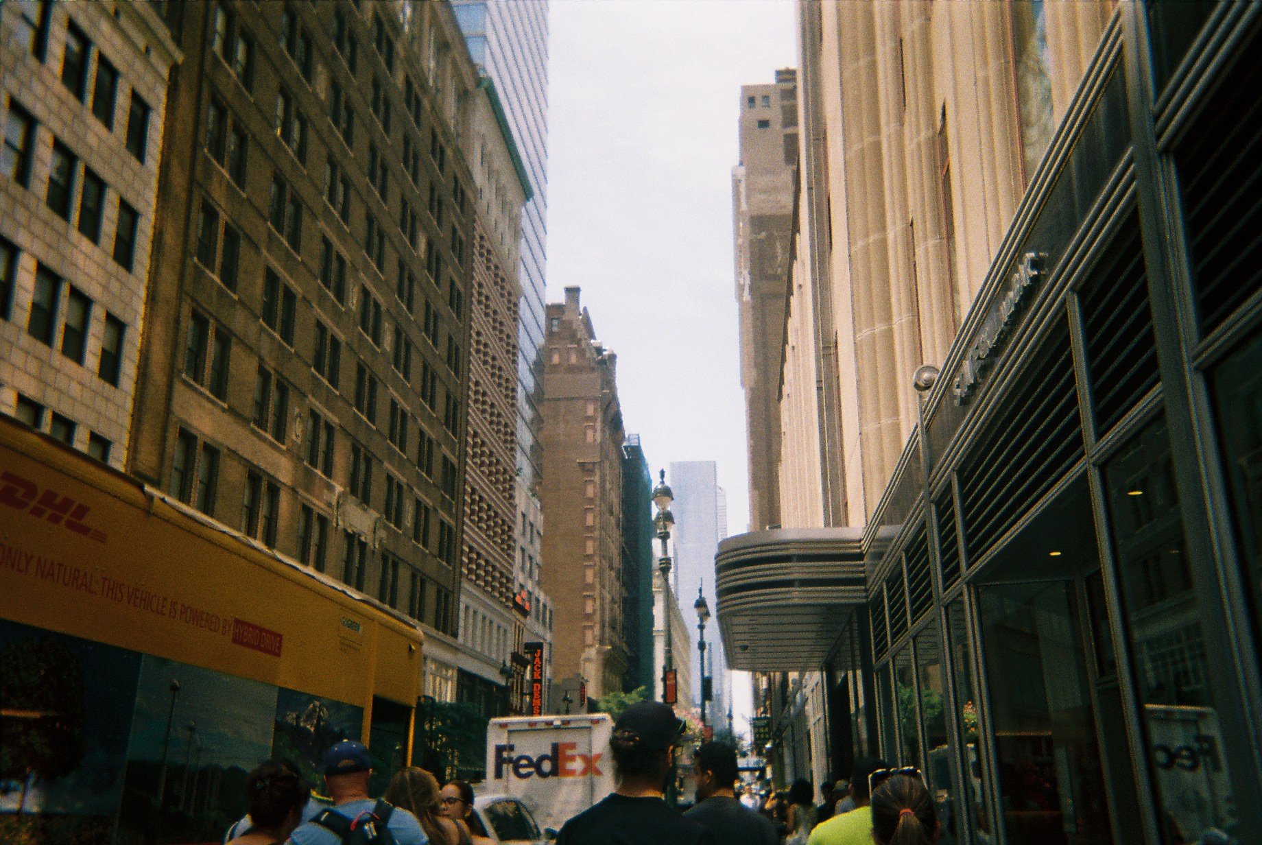 Crowd of people walking on a busy city street surrounded by tall buildings, with a FedEx truck and a yellow DHL vehicle visible.