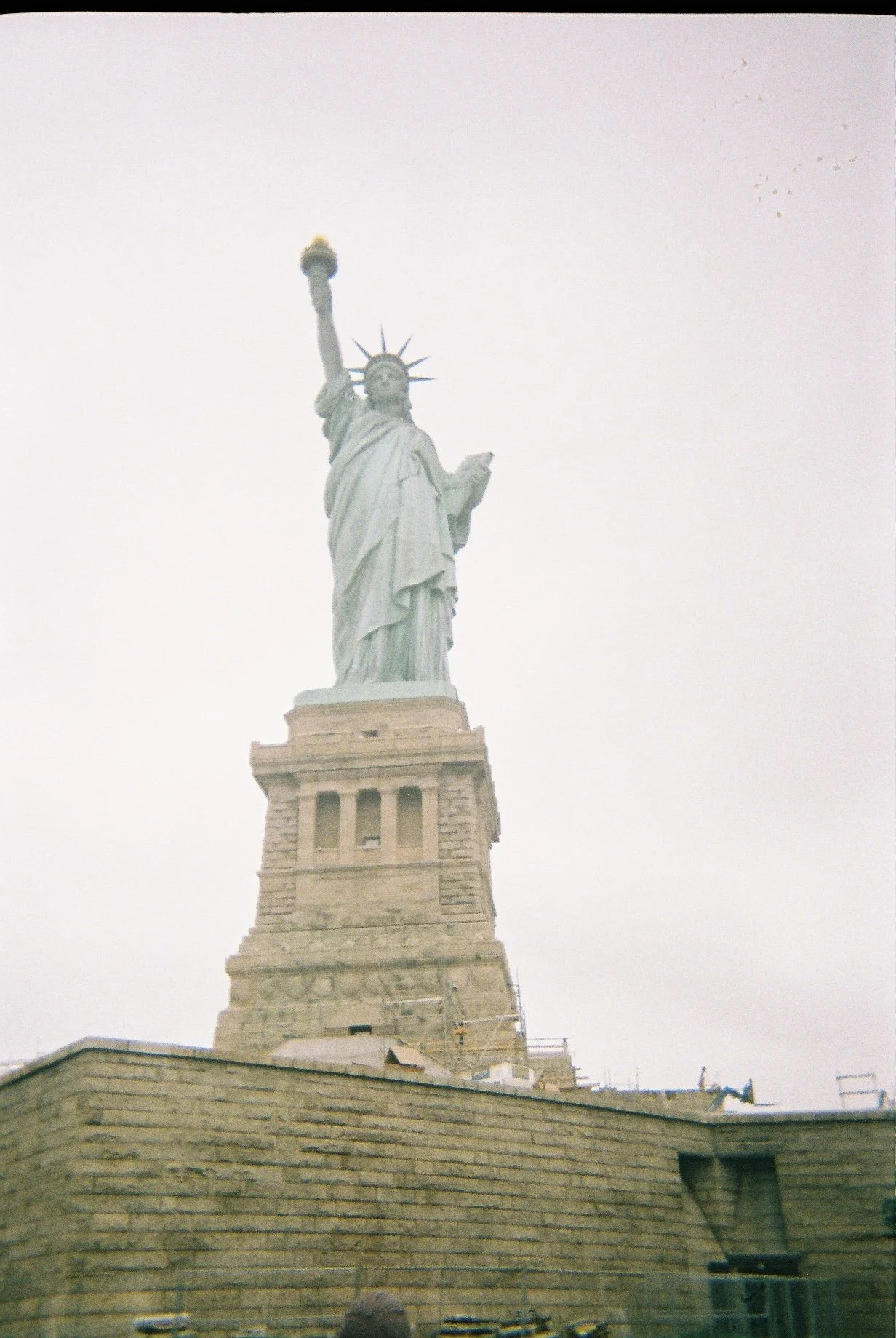 Photograph of the Statue of Liberty on Liberty Island, showing the statue's front with the torch held high in her right hand and the tablet in her left hand. The base of the statue is visible with surrounding customs buildings.