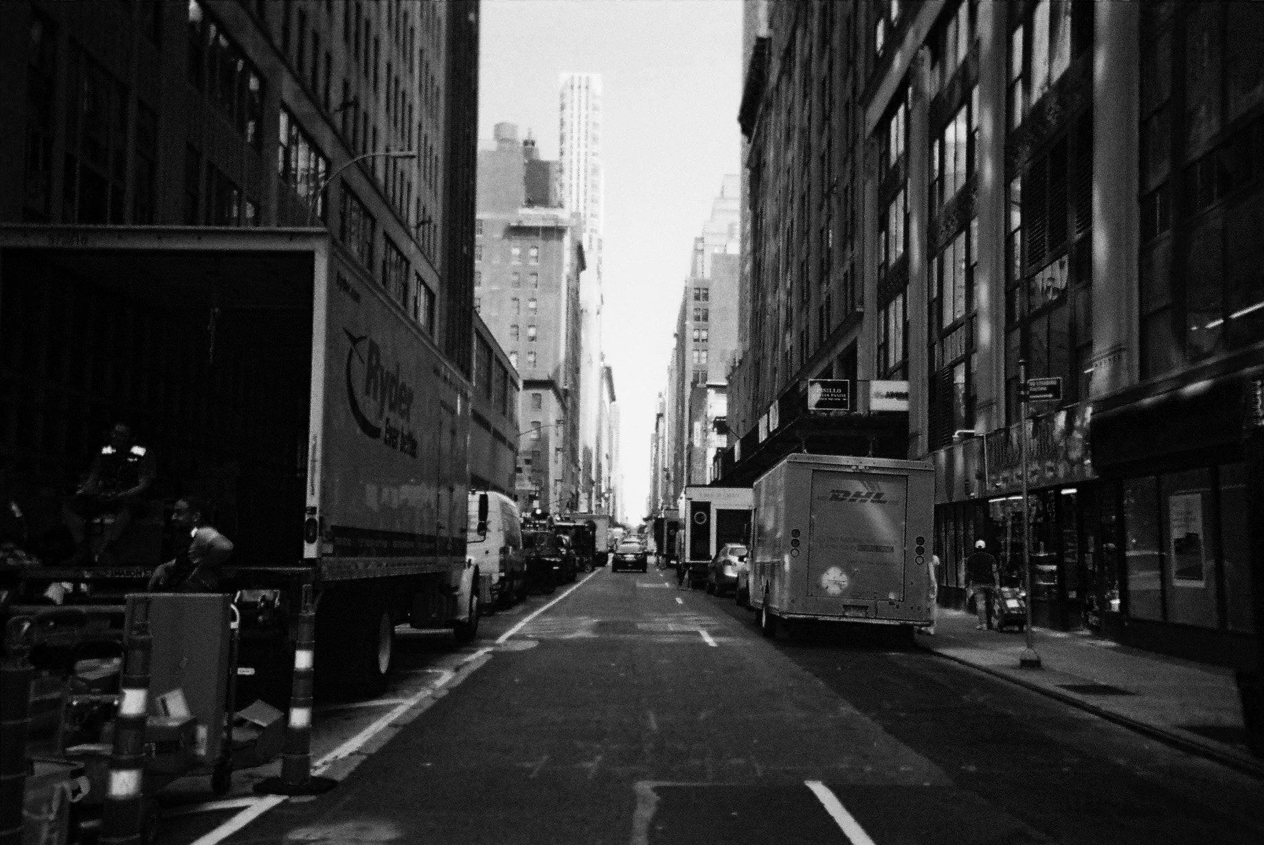 A black and white photo of a city street with cars, trucks, and buildings on both sides. Tall skyscrapers are visible in the background.