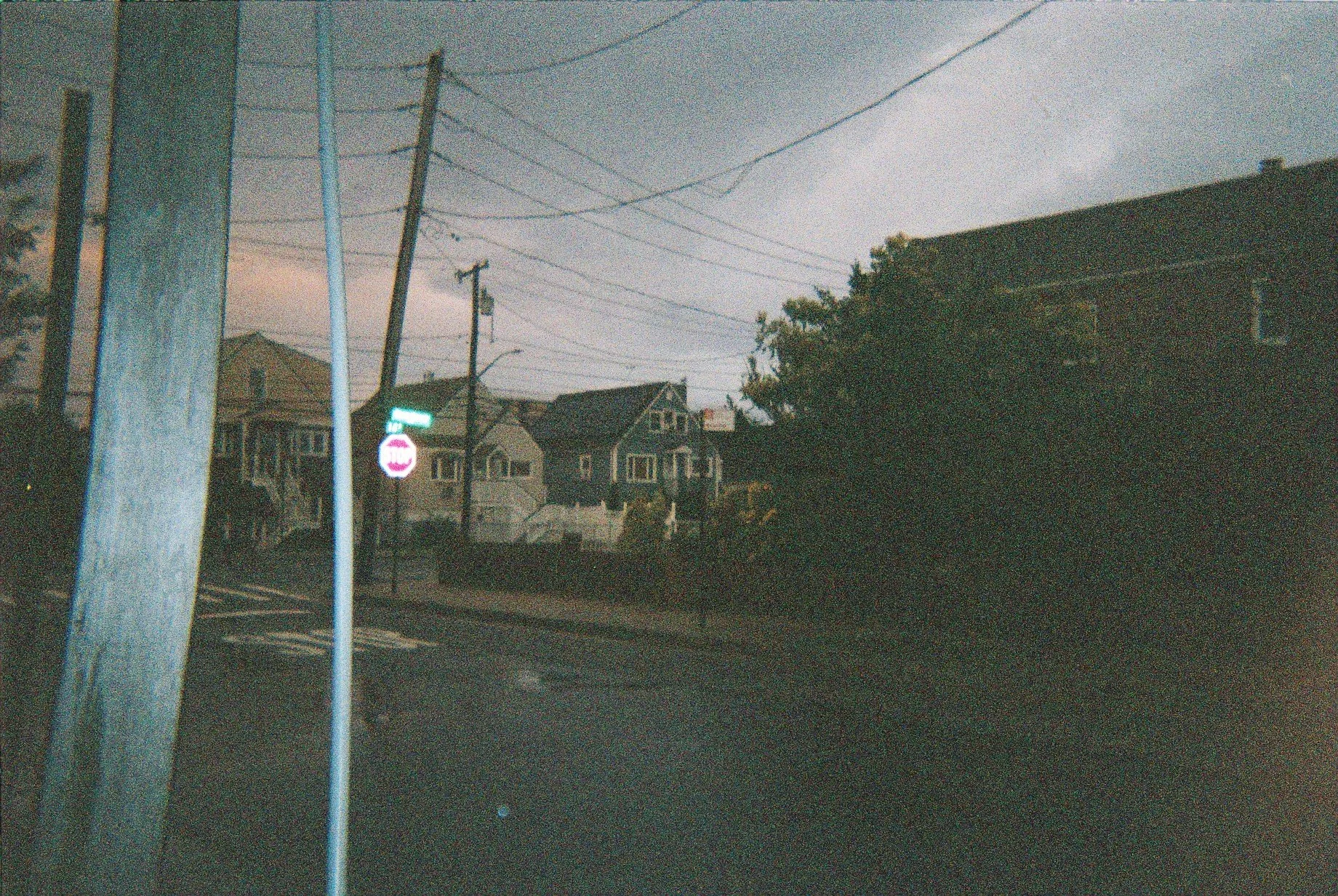 Residential street scene during dusk with utility poles, street sign, houses, and dark cloudy sky.