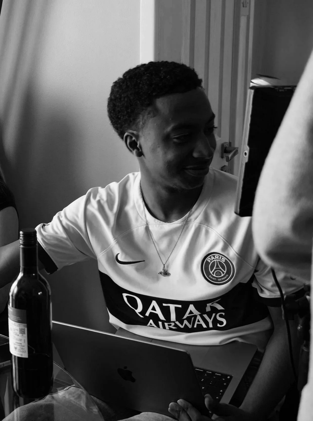 Young man with short curly hair wearing a Paris Saint-Germain soccer jersey, sitting at a table with an open laptop, a bottle, and a person holding a device in front of him, in a room with a door and curtains.
