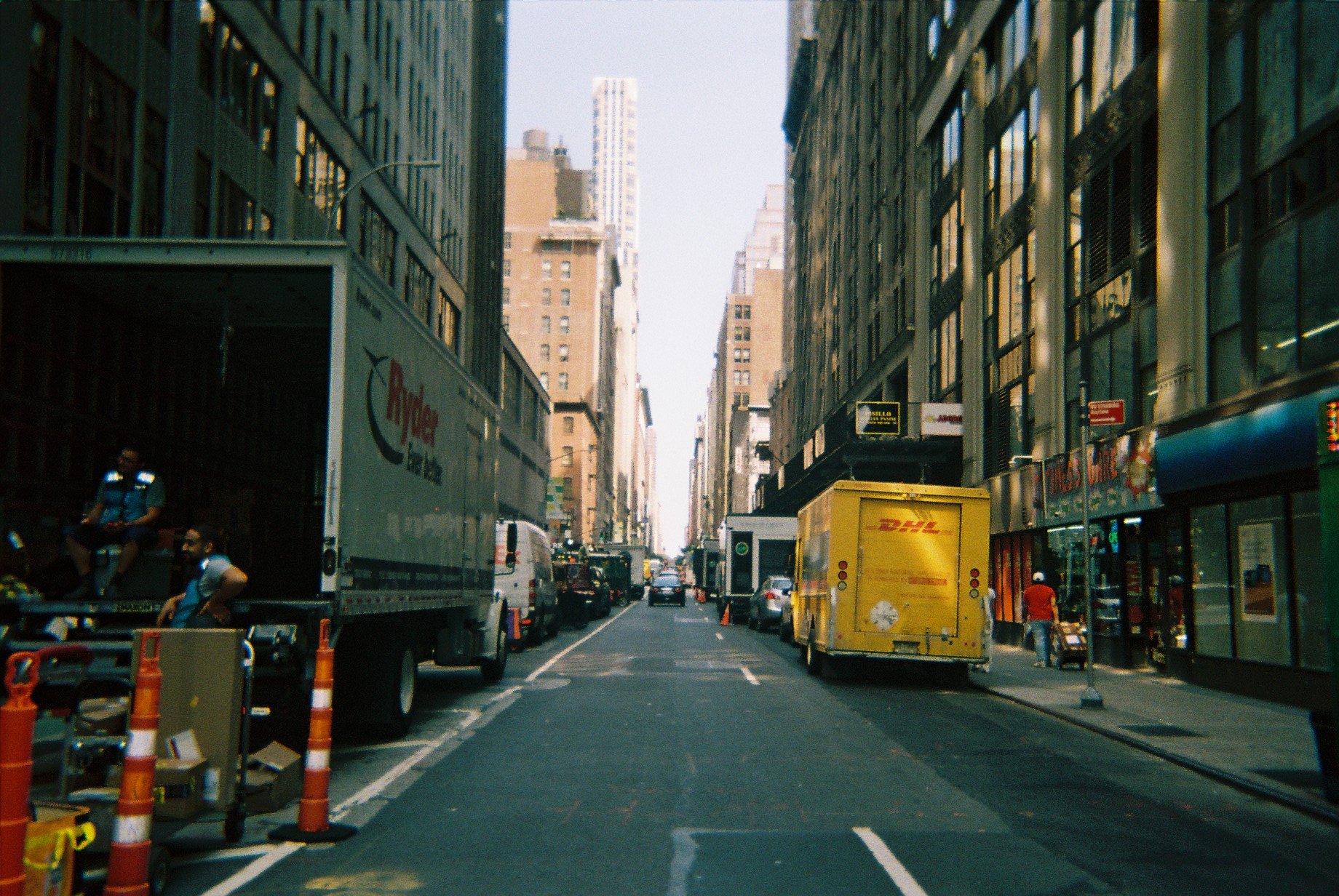 City street scene with parked delivery trucks and cars, tall buildings on either side, pedestrians, and construction cones on the road.