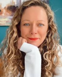 A woman with long, wavy brown hair and light makeup, wearing hoop earrings and a beige top, posed against a red background.