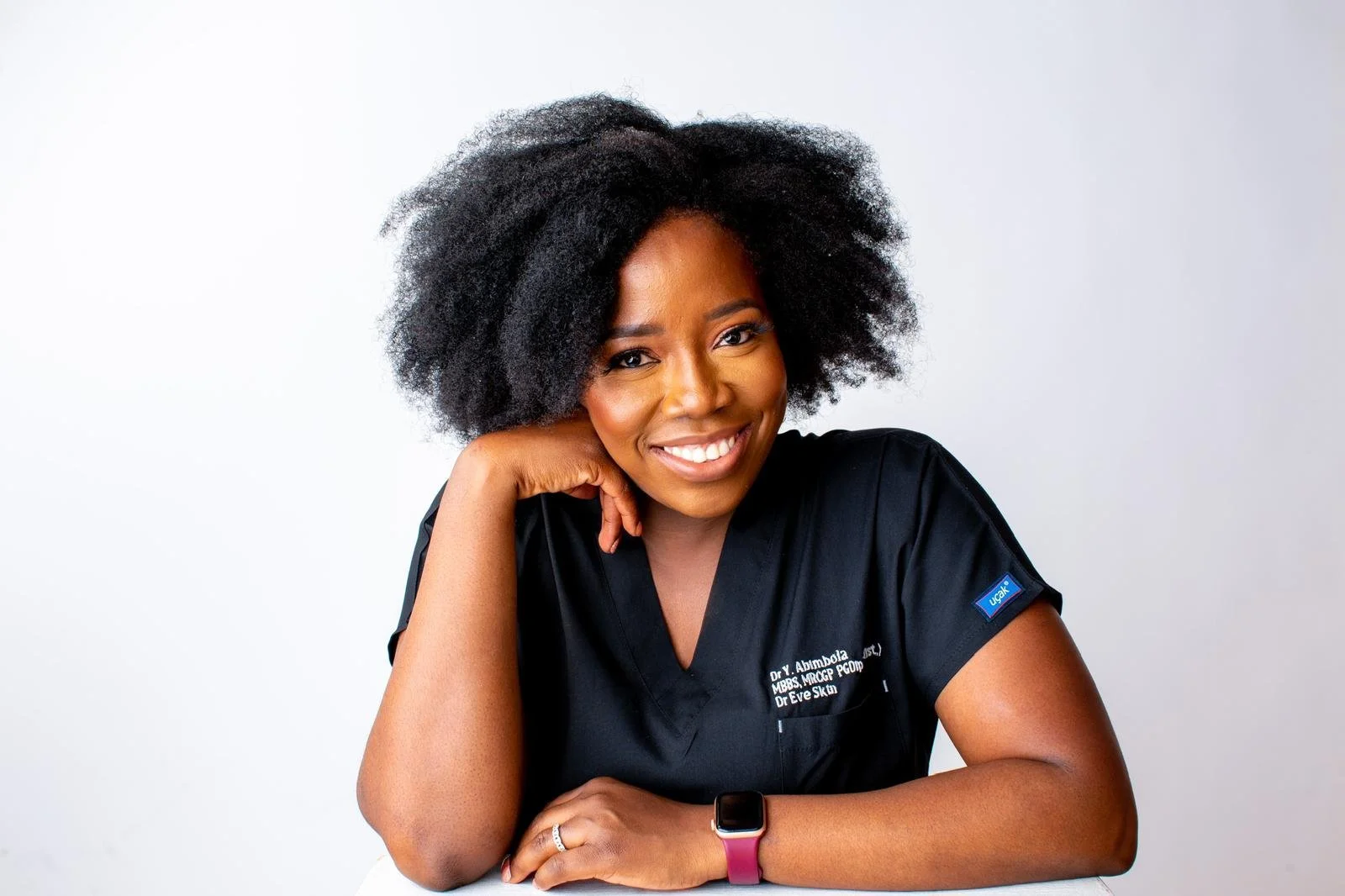 A woman with long, wavy brown hair and light makeup, wearing hoop earrings and a beige top, posed against a red background.