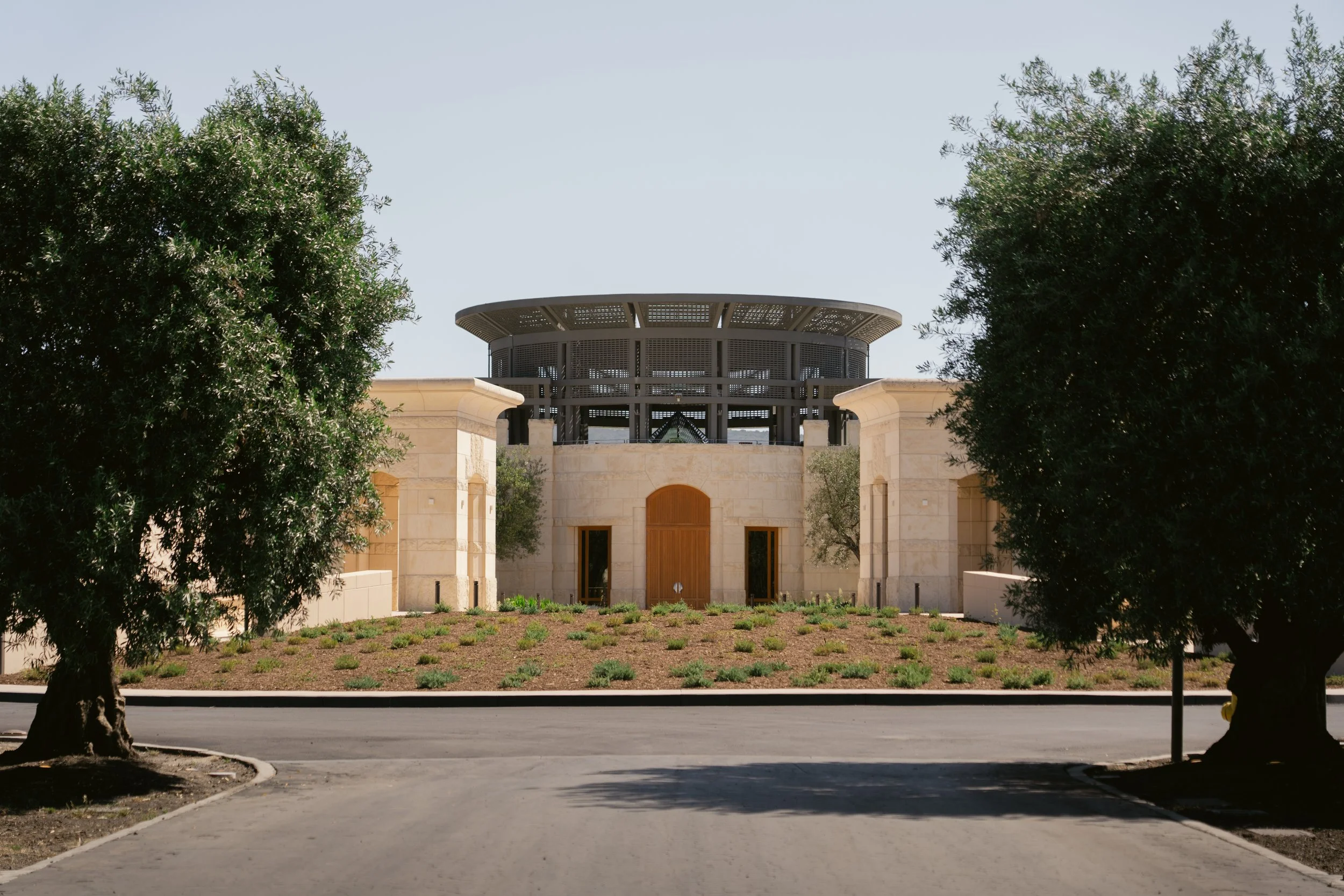A modern building with a rounded top, beige exterior stone walls, wooden doors, and a landscaped front yard, framed by two large trees. Opus One Winery in Napa.