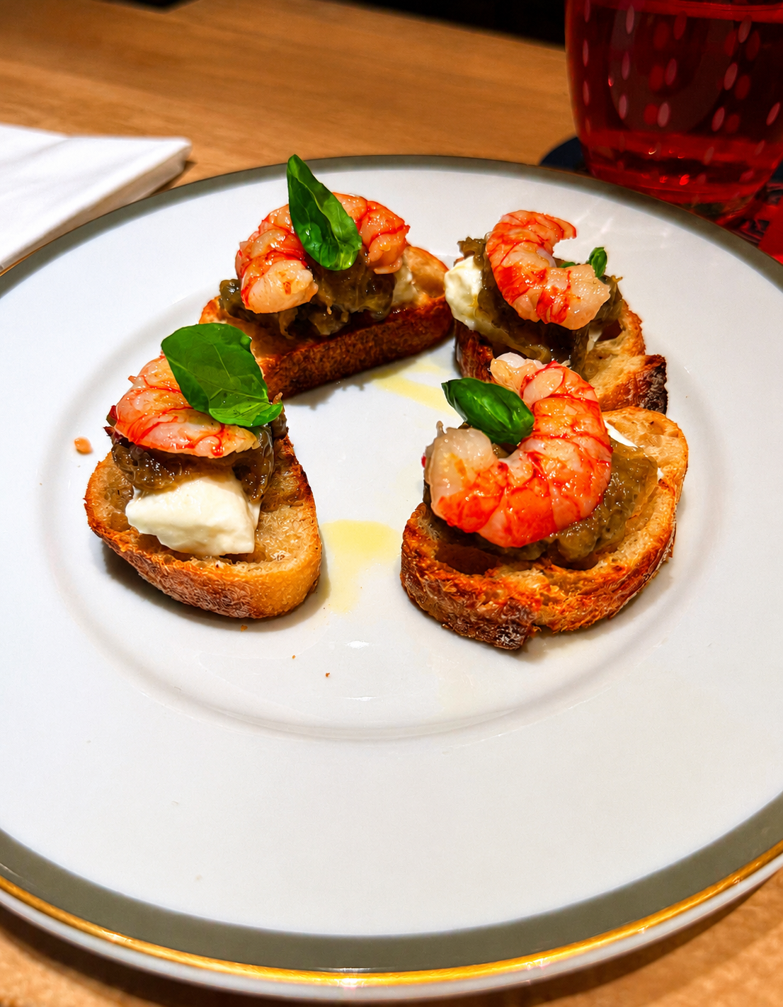 Three pieces of toasted bread topped with shrimp, a dollop of white cream, green basil leaves, and a dark green spread, arranged in a circle on a white plate.  Langosteria in Paris.