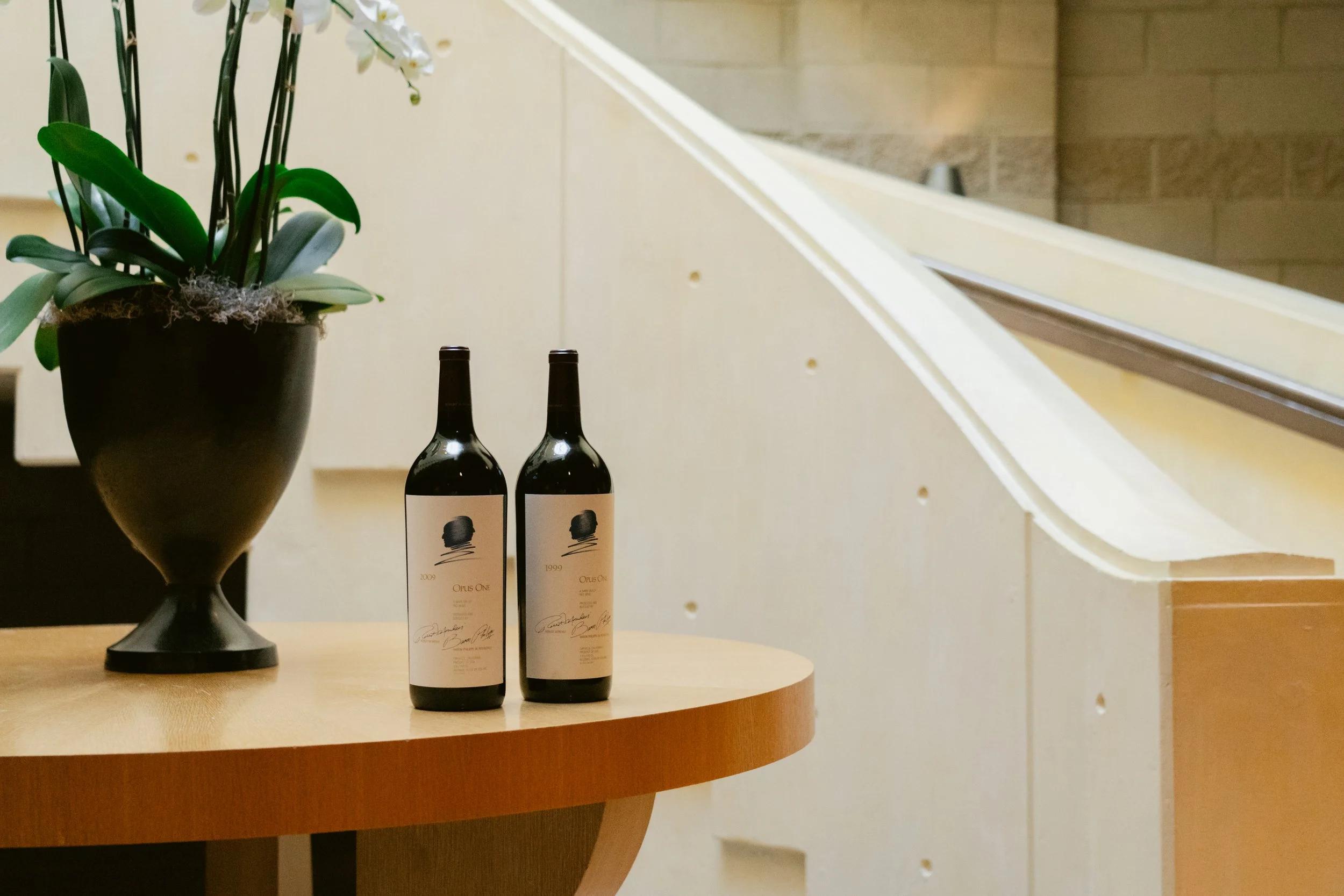 A black vase with white orchids, two bottles of wine with cream labels, and a wooden table surface in a room with beige walls. Opus One Winery in Napa.