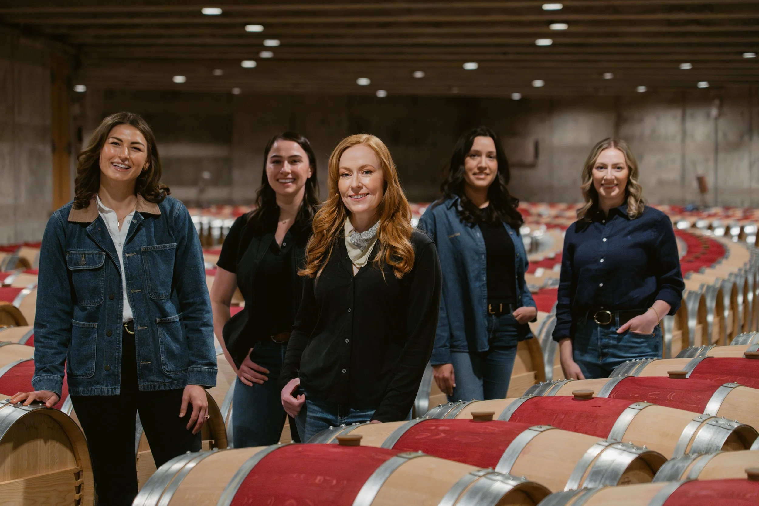 Five women standing in a wine cellar with wine barrels, smiling at the camera. Opus One Winery in Napa.