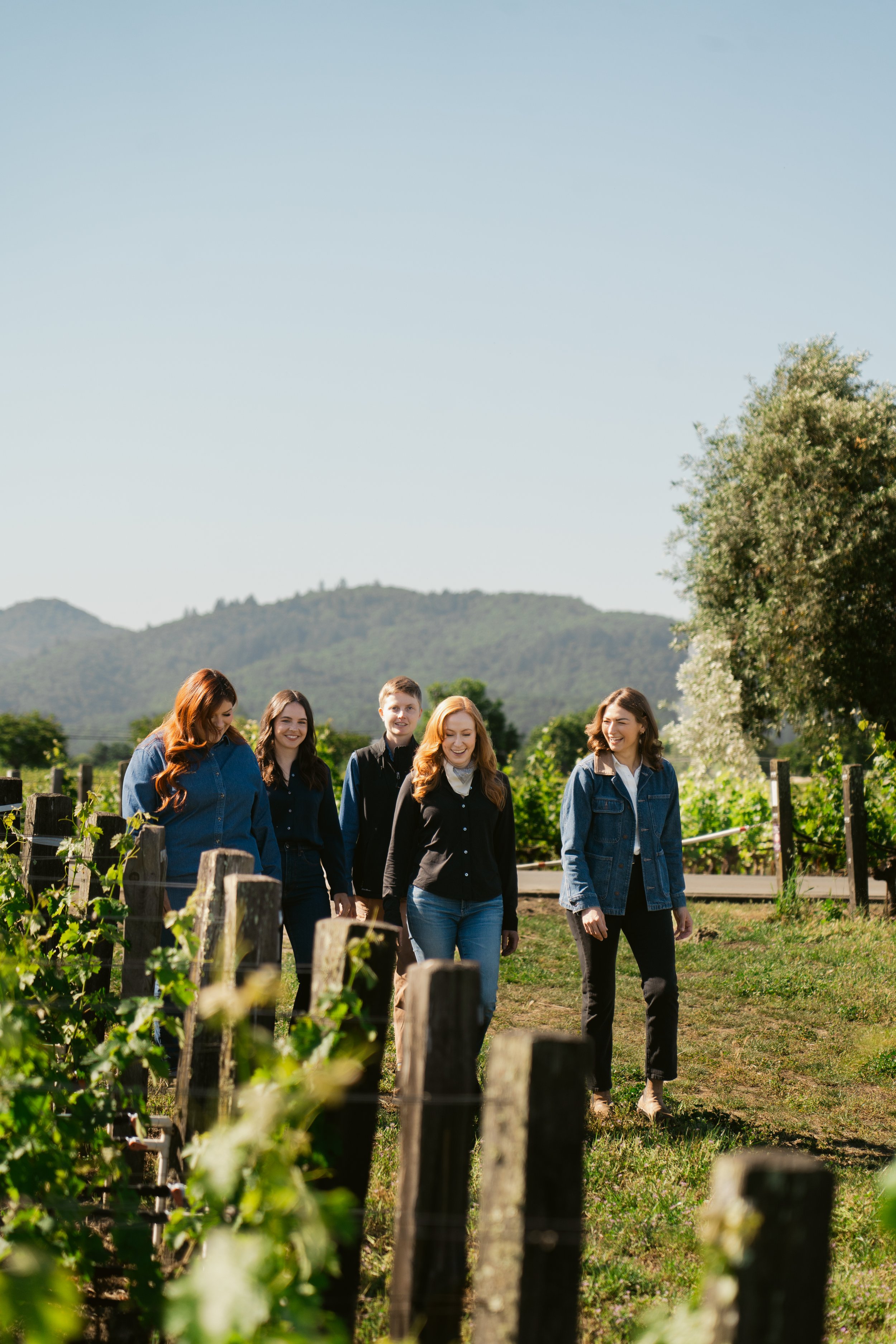 A group of five women walking through a vineyard on a sunny day, smiling and talking with each other. There are hills and trees in the background. Opus One Winery in Napa.