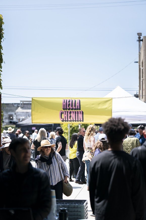 Crowd of people gathered outdoors at a festival or market with a yellow banner reading 'HELLO CHENIN' in pink and black letters, and white tents in the background.