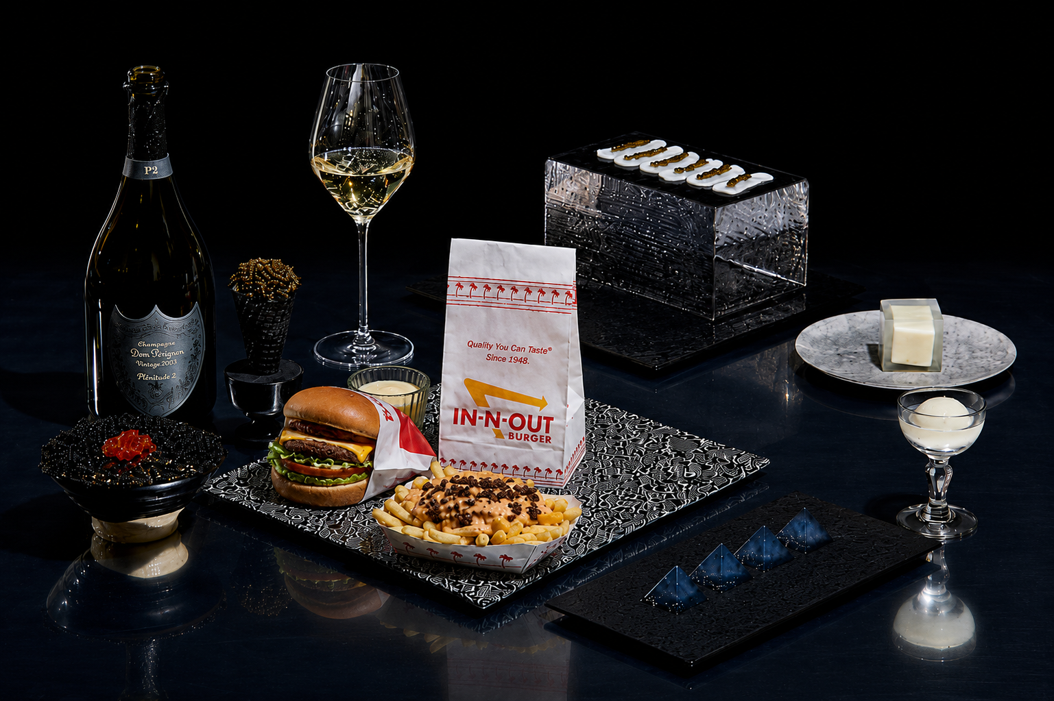 A black table set with a bottle of champagne, a glass of white wine, a burger wrapped in branded paper, a container of mac and cheese with toppings, a slice of white chocolate on a plate, a small bowl of caviar, a glass of white sauce, a black tray with chocolates and a QR code, and a box of chocolates.