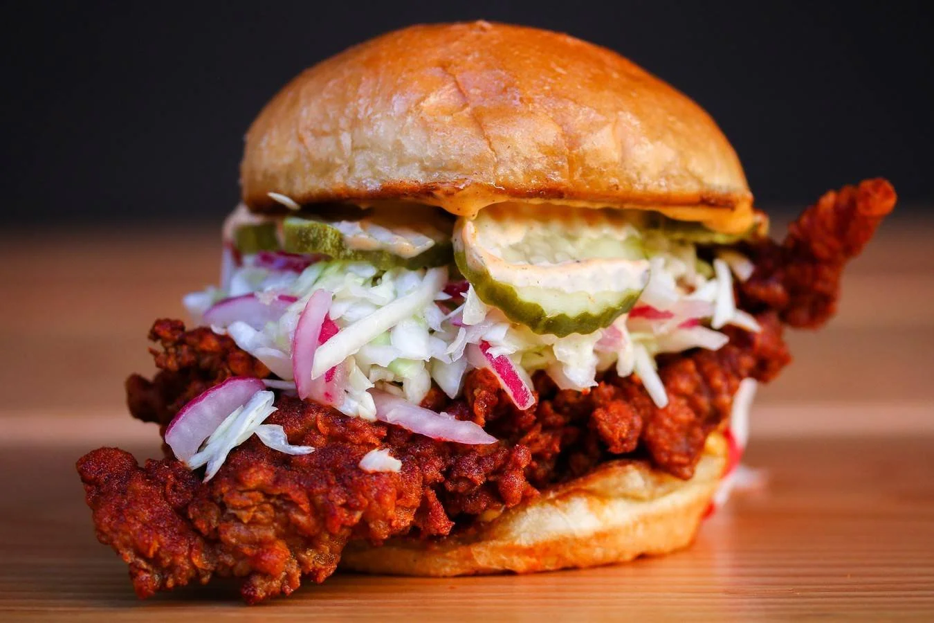 Close-up of a fried chicken sandwich at Howlin' Rays with coleslaw, pickle slices, and a soft bun, on a wooden surface with a dark background.