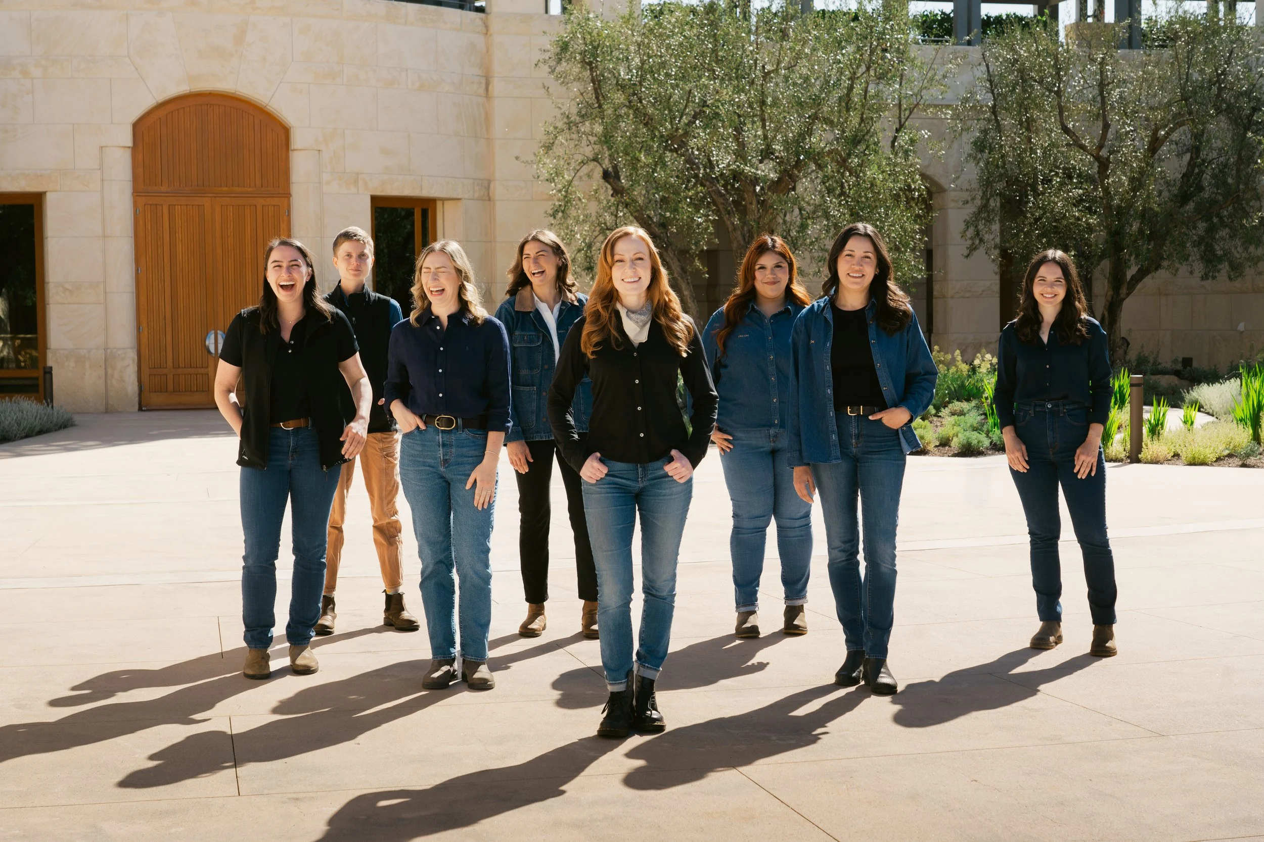 A group of eight young women walking outdoors on a sunny day, smiling and enjoying each other's company, with greenery and a modern building in the background at Opus One winery in Napa.
