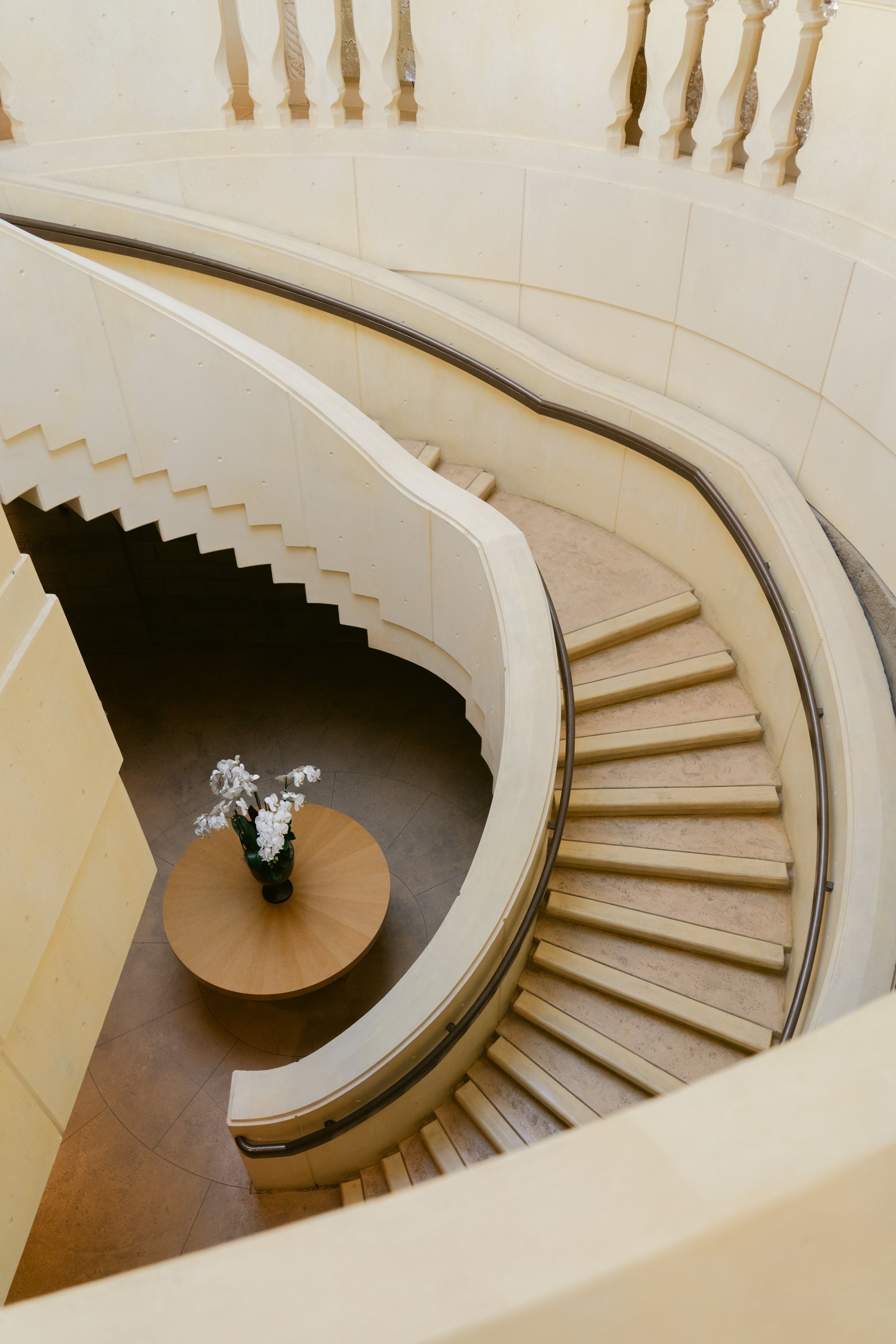 A spiral staircase with cream-colored walls and beige steps, decorated with a dark handrail. At the bottom, there is a round wooden table with a black vase holding white flowers. Opus One Winery in Napa.