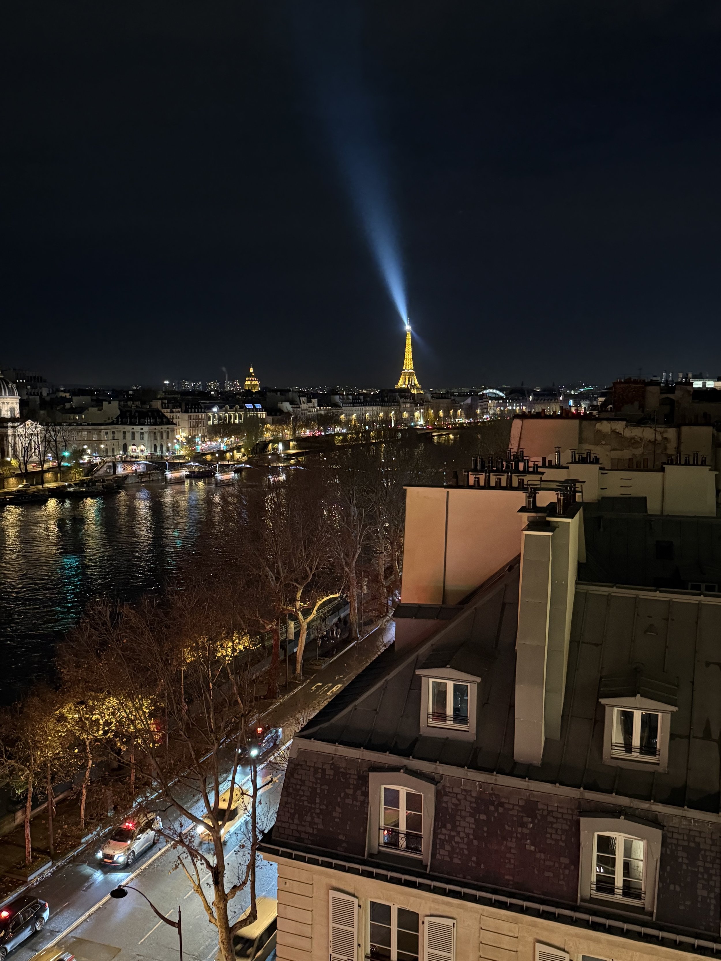 Nighttime city scene with buildings, a river, and a tall tower with bright lights and a spotlight beam shooting upward.