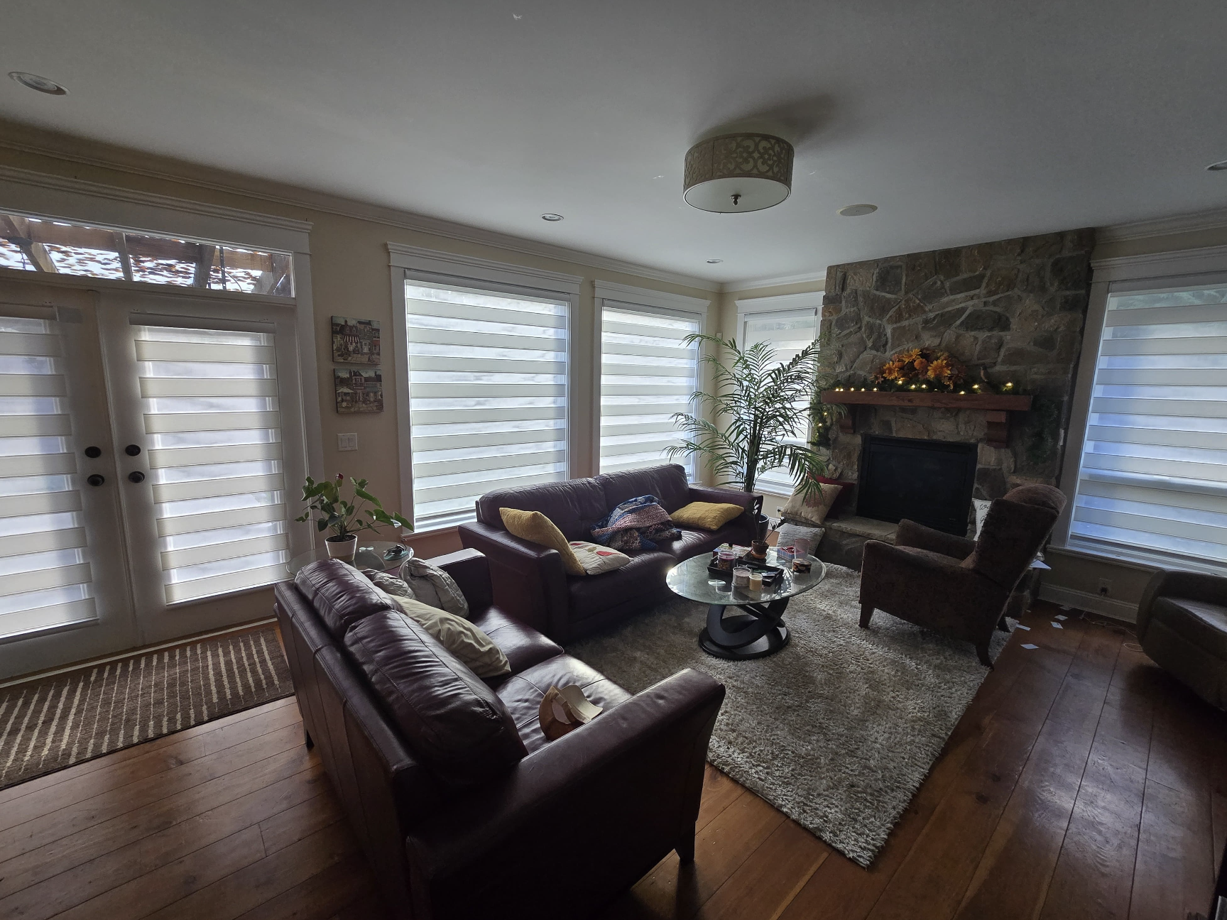 Living room with leather sofas, a stone fireplace decorated with flowers and lights, large windows with striped blinds, and wooden flooring.