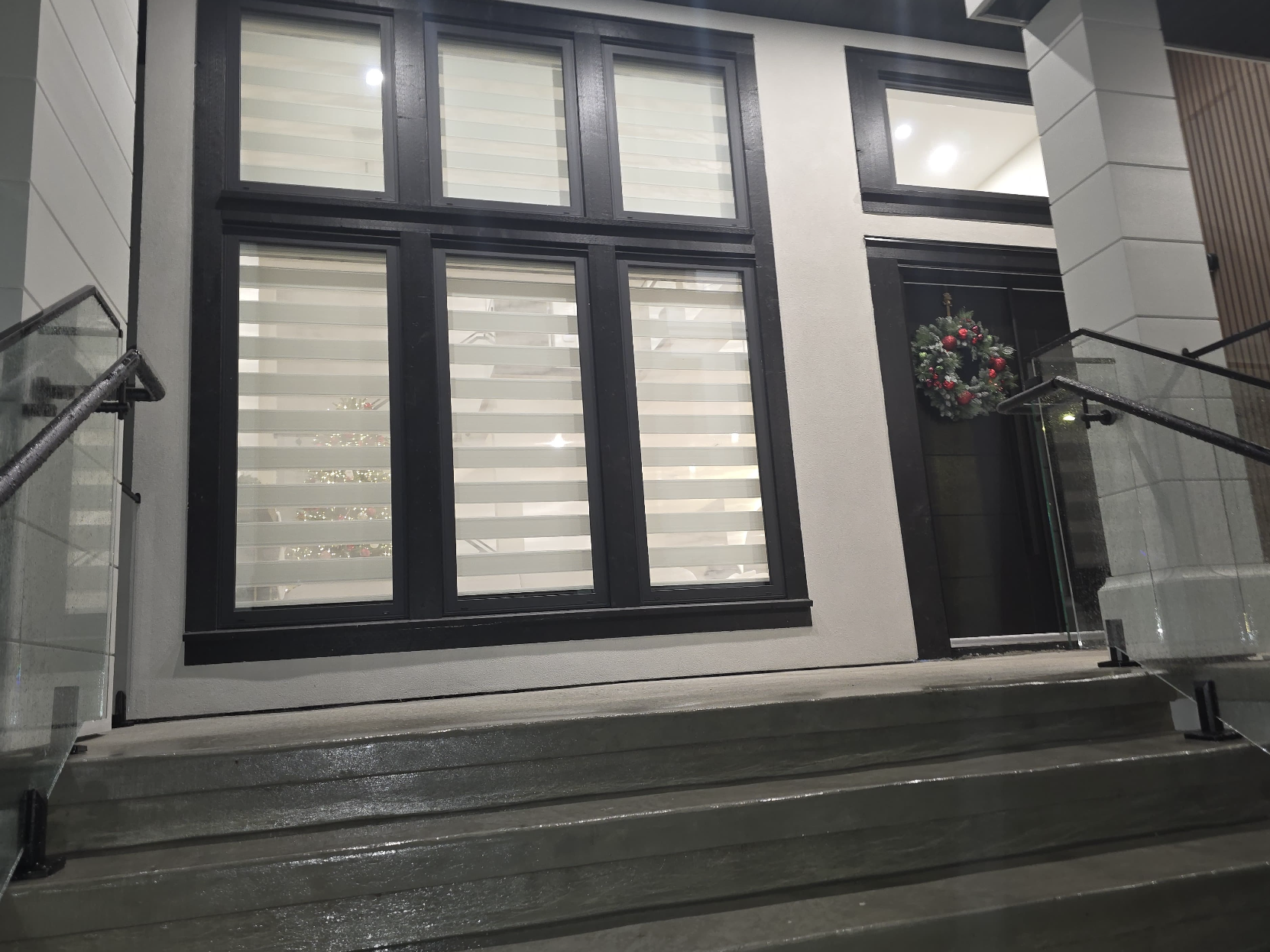 The entrance of a modern house with stairs leading up to a glass door decorated for Christmas with a wreath; large window with blinds on the front of the house.