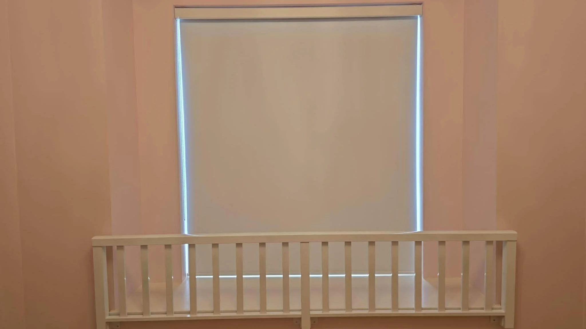A nursery room with pink walls, a closed window with a beige roller shade, and a white wooden crib in front of the window.