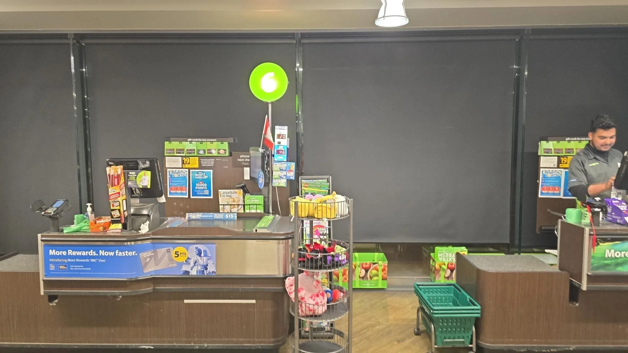 Grocery store checkout counter with a worker on the right, shelves with snacks and toys in front, black wall in the background, and various signs and promotional materials on the counter.