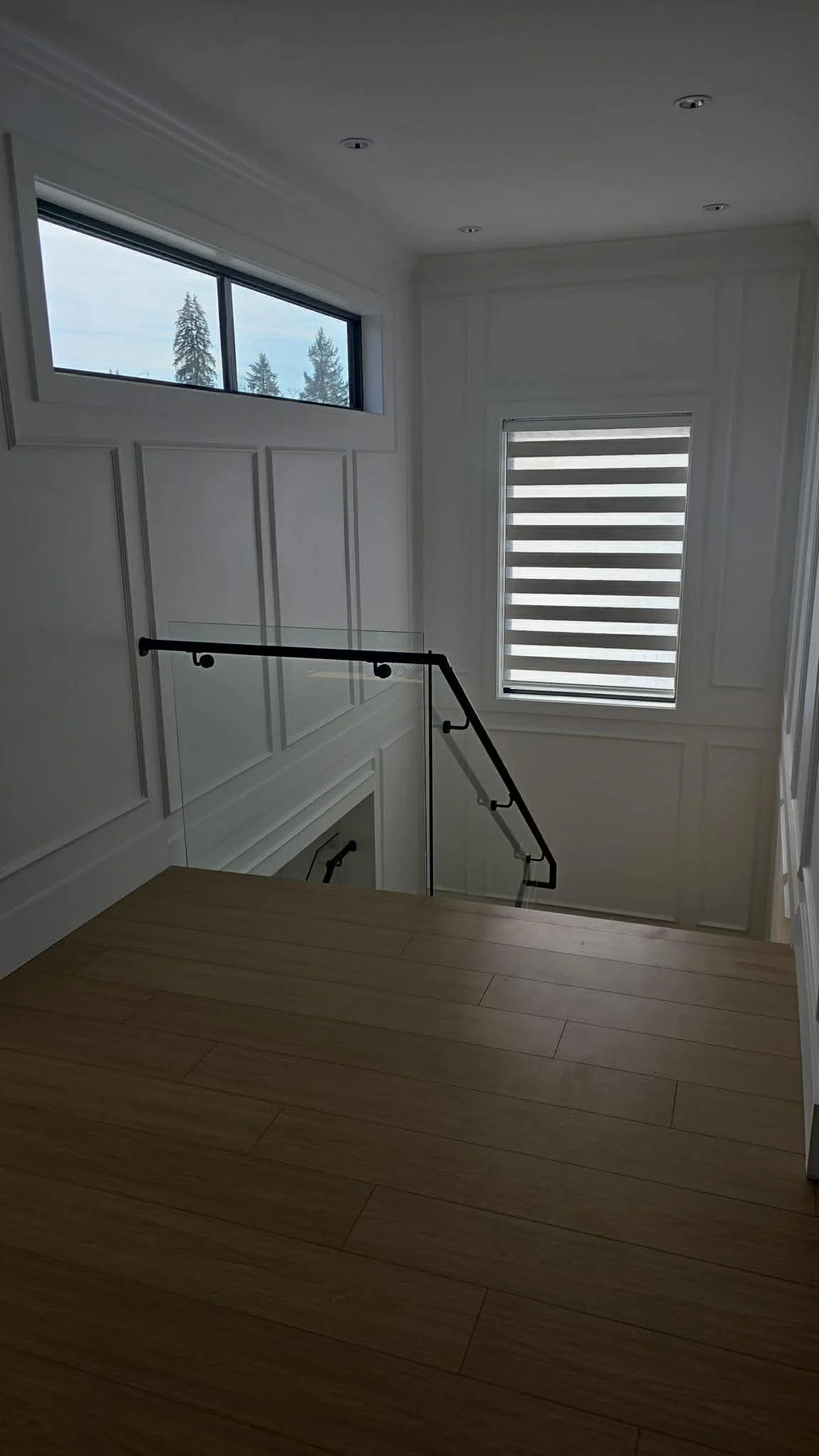 Interior view of a staircase area with white paneled walls, a black handrail, and windows with a view of trees outside. Light streams through the windows, illuminating a wooden floor.