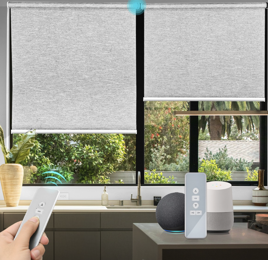 View of a kitchen with two smart blinds partially lowered, a hand holding a remote control, and smart home devices including a smart speaker and smart display on the kitchen counter.