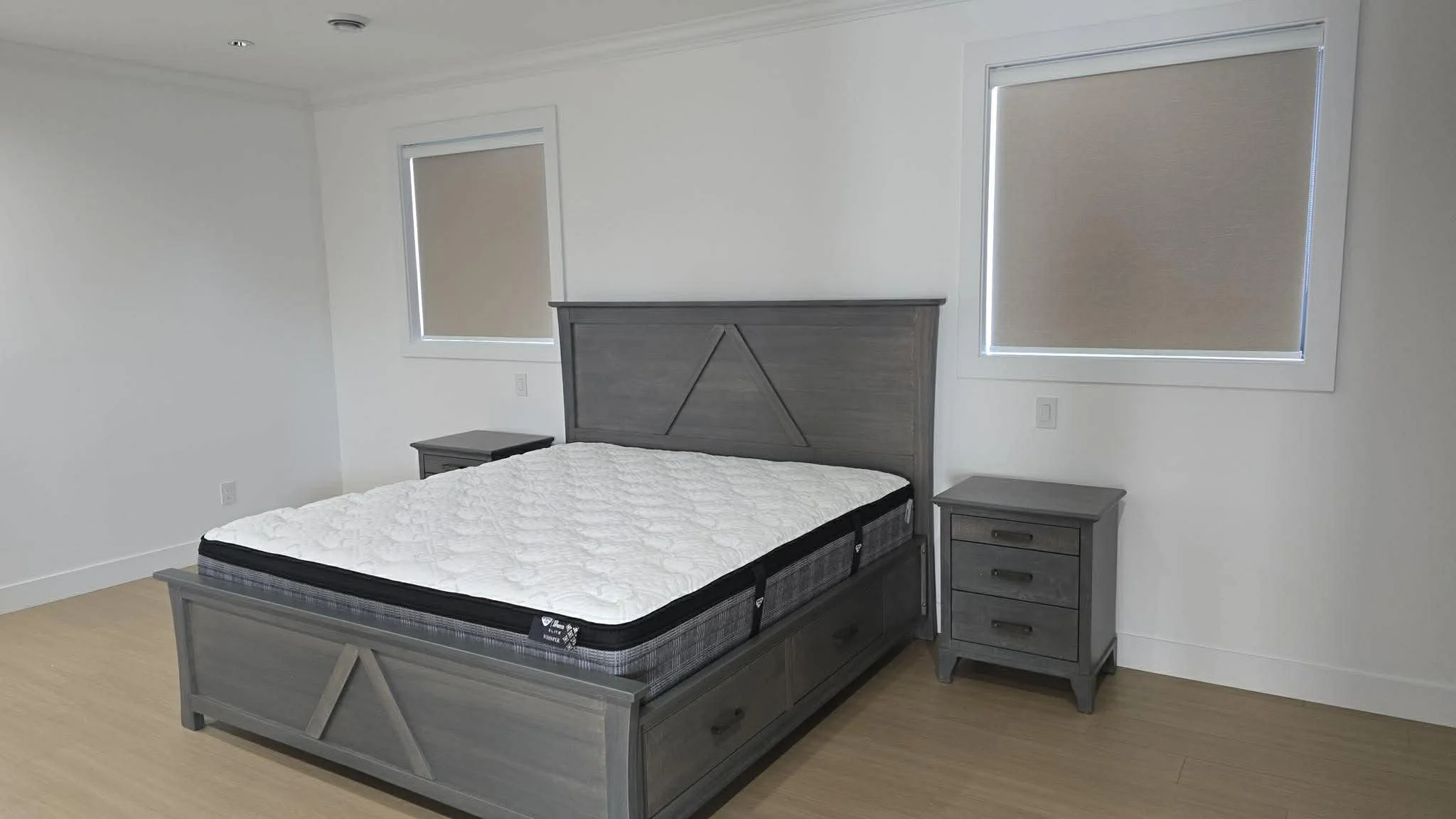 A bedroom with a wooden bed frame and a mattress. There are two small wooden nightstands, one on each side of the bed. The walls are white, and there are two windows with blinds, one on each side of the bed.