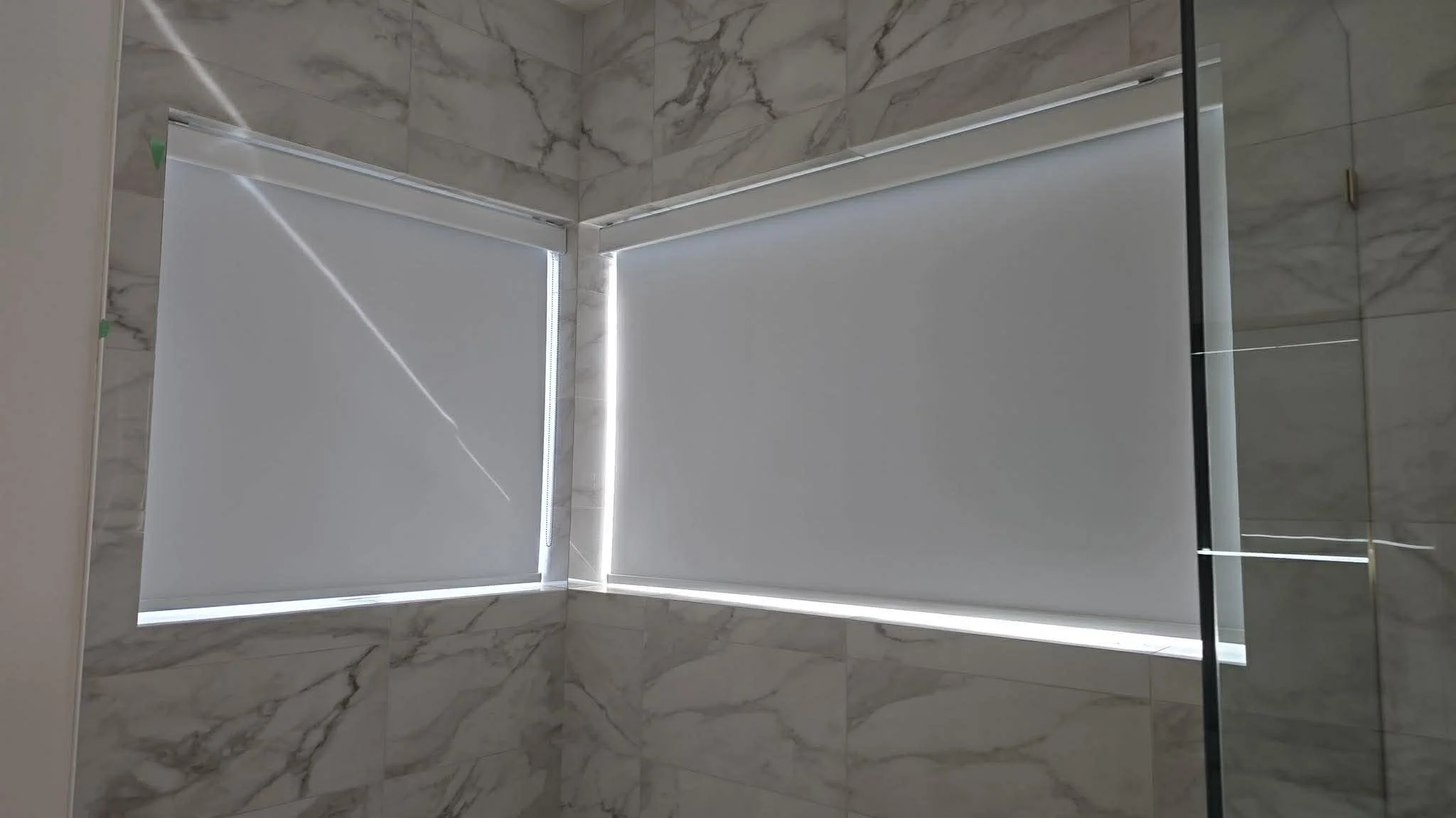 Bathroom with two windows covered by white roller blinds, marble tiled walls, and a glass shower panel on the right side.
