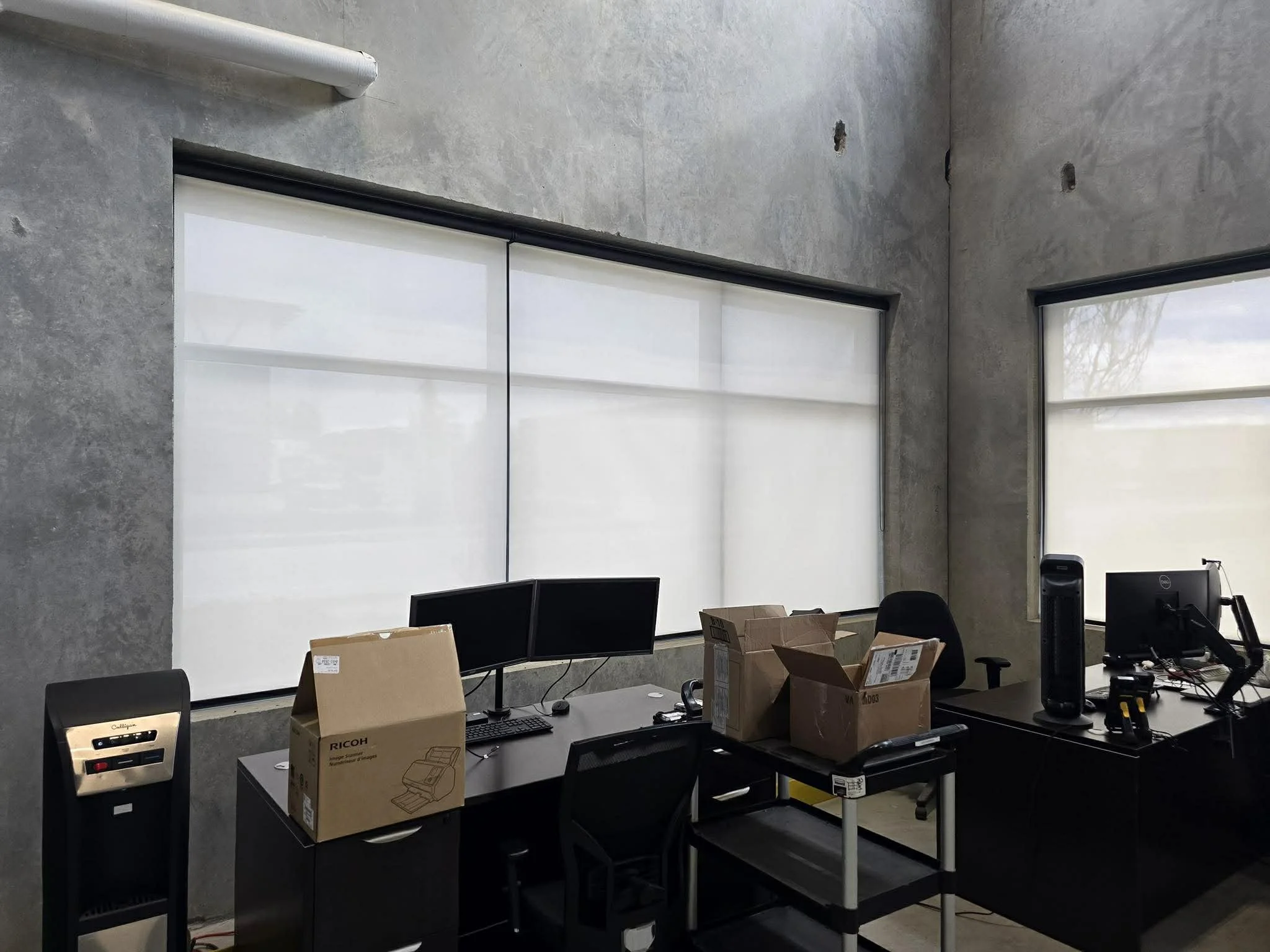 Office interior with two large horizontal windows covered with white blinds, concrete walls, black desks with monitors, cardboard boxes on desks, and office chairs.