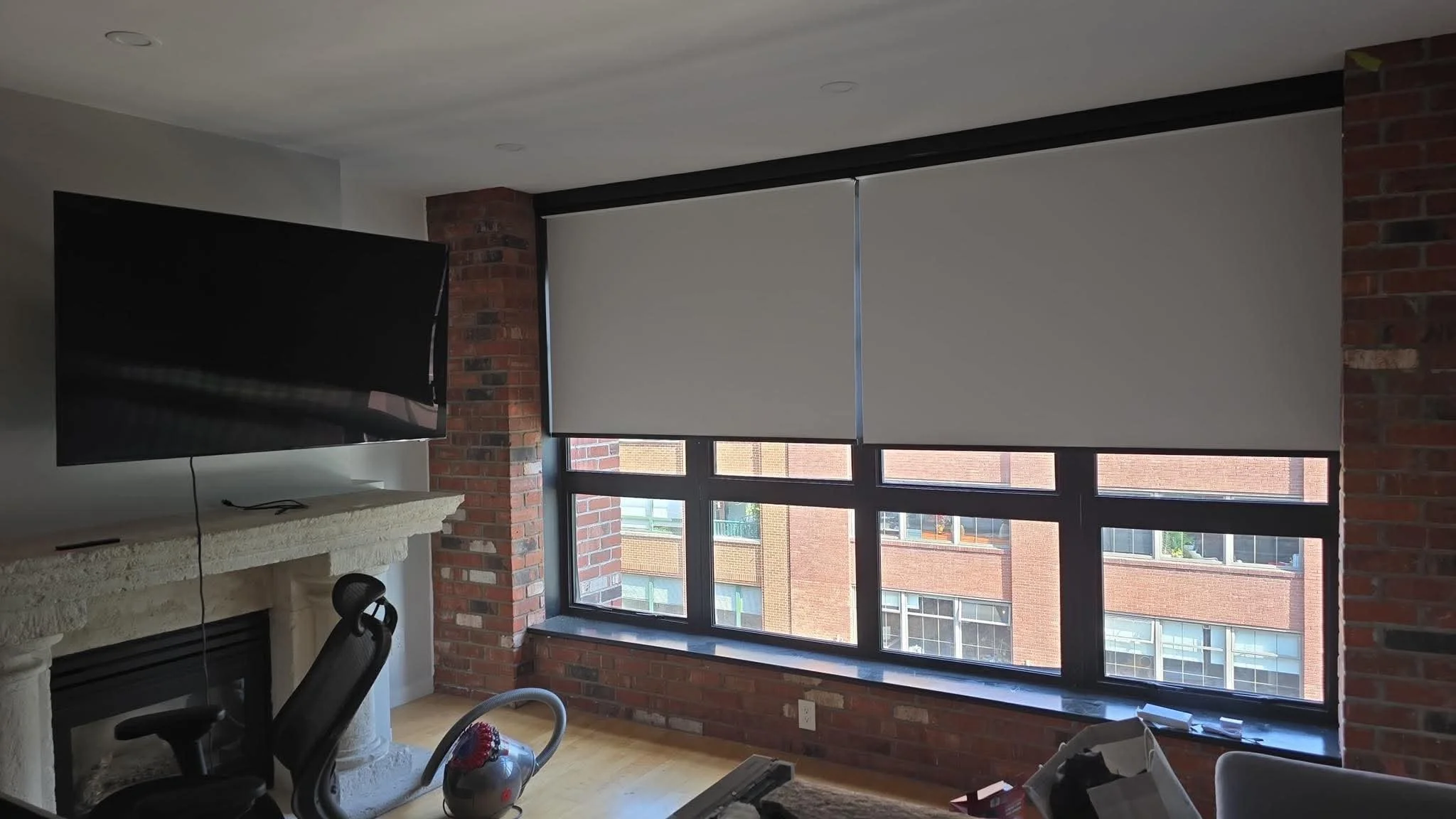 Interior view of a living room with large windows covered by white blinds, brick walls, a flat-screen TV mounted on a stone fireplace mantel, and some furniture and items on the floor.