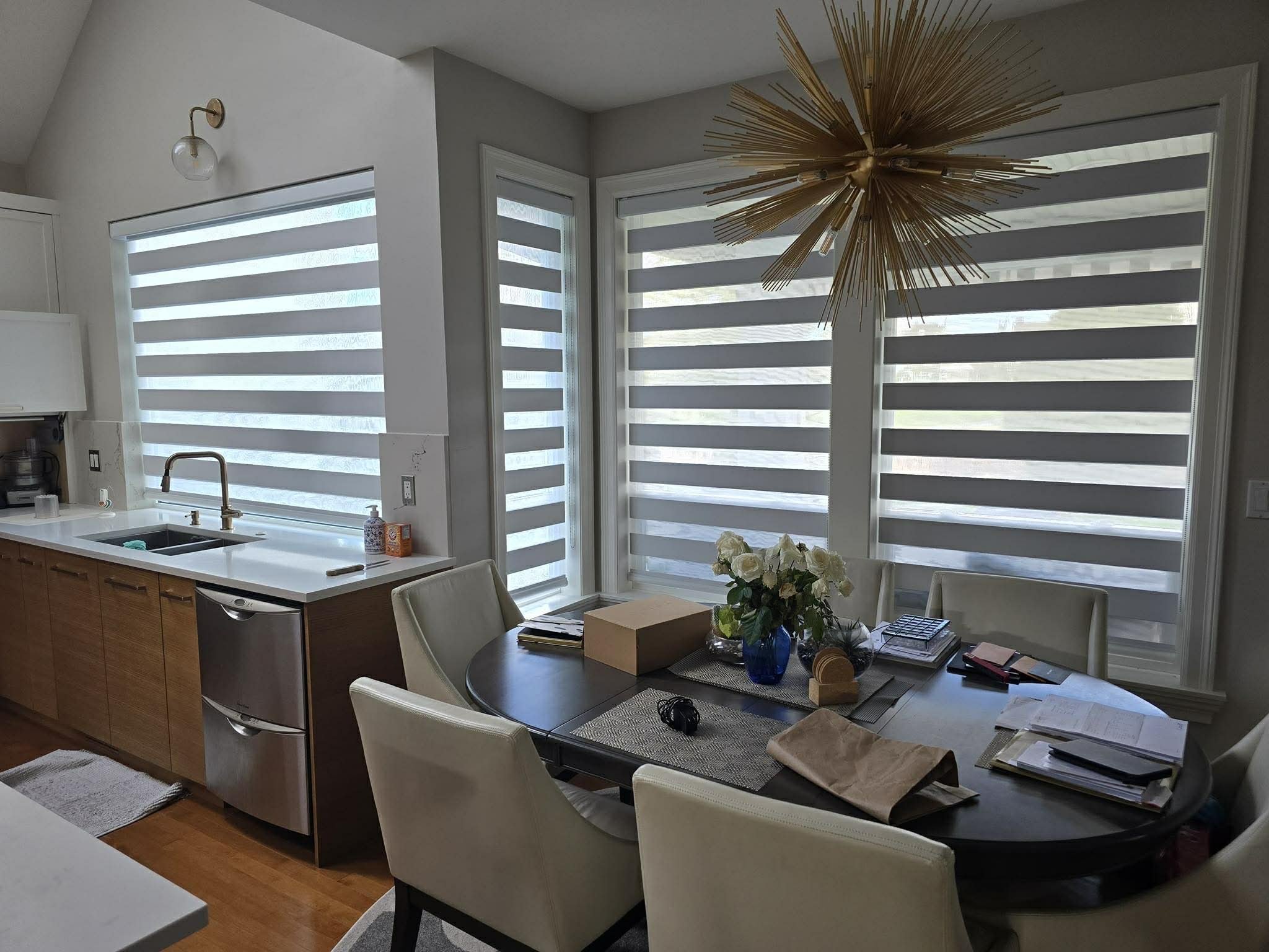 Dining room with large windows covered in zebra blinds, dark oval table with beige chairs, a vase with white flowers, and various scattered papers and objects. Kitchen area with sink, faucet, and white countertop visible in the background. Gold starburst chandelier hangs above the table.