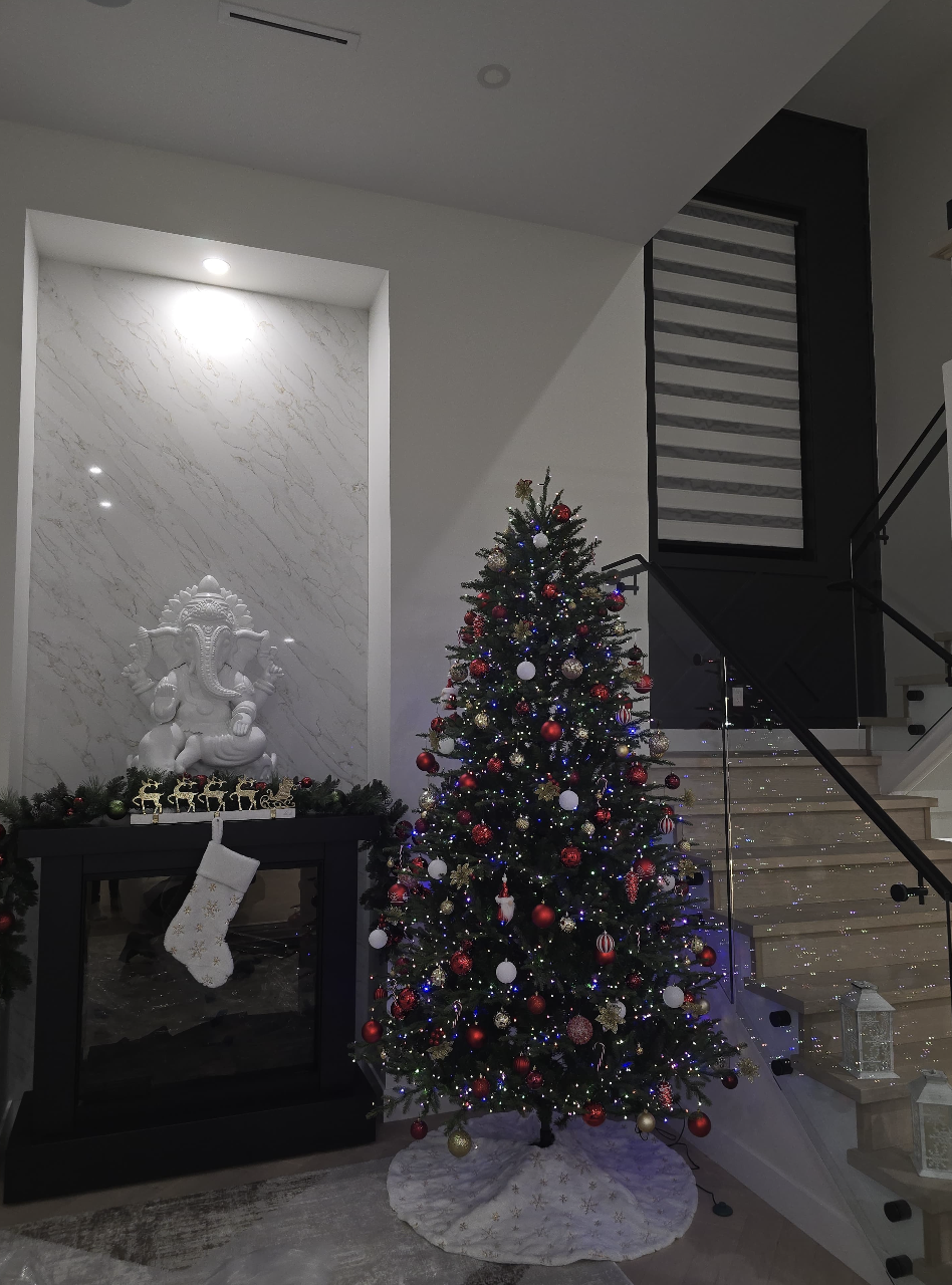 Decorated Christmas tree with red, gold, and white ornaments, lights, and a white tree skirt, positioned in a modern home interior near stairs and a fireplace with a white Ganesha statue and Christmas stocking.