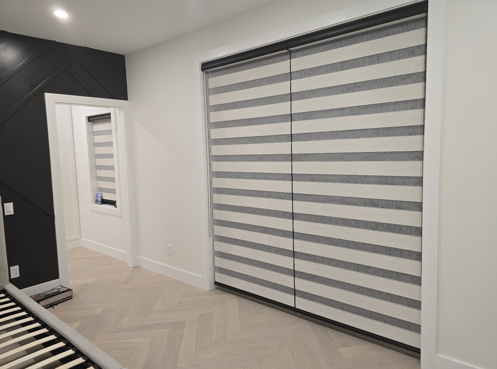 Interior room with light-colored hardwood floor, black and white walls, a large window with striped shades, a small window with striped shades, and a partial view of a bed frame.