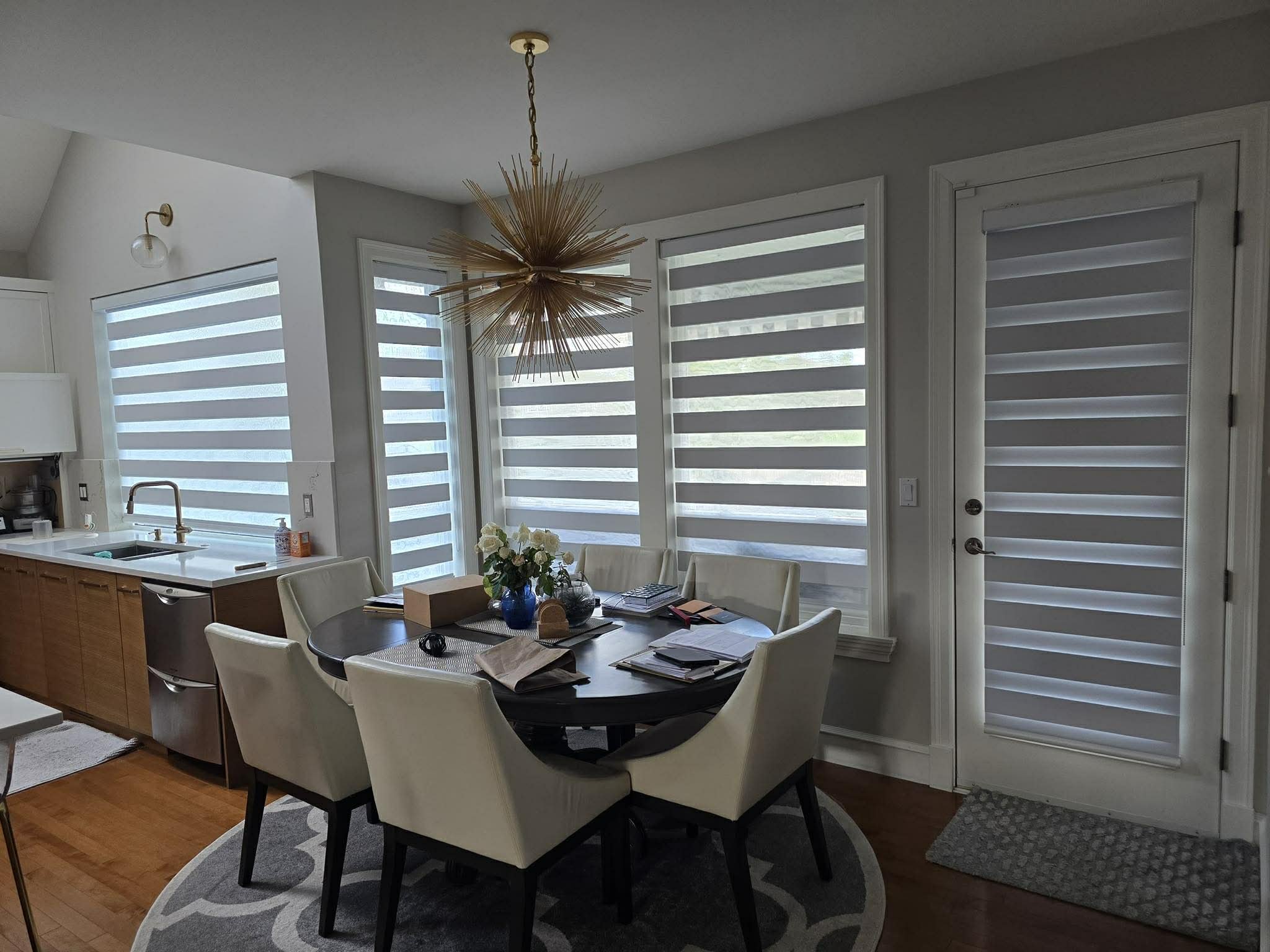 Dining area with a round black table, surrounded by six white chairs, in front of large windows with zebra shades. The table has papers, a box, and a flower arrangement. A pendant light hangs above.