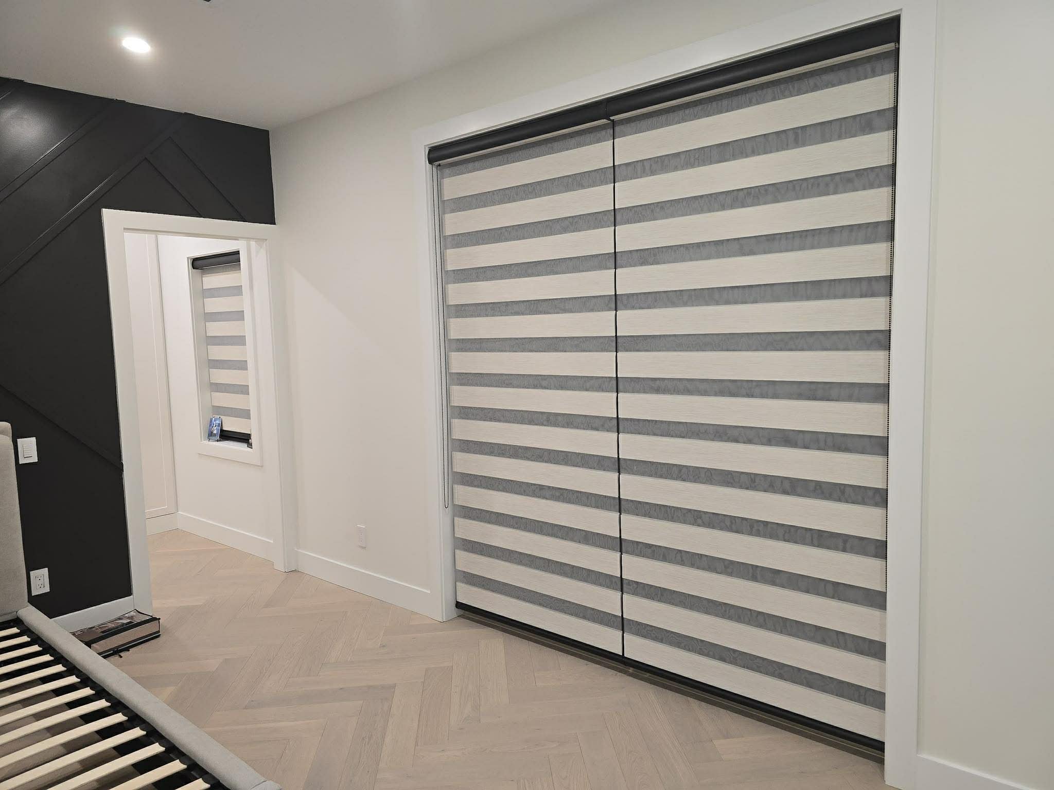 Interior of a bedroom with white and black walls, a window with a striped roller shade, a light-colored herringbone wood floor, and a wardrobe with striped sliding doors.