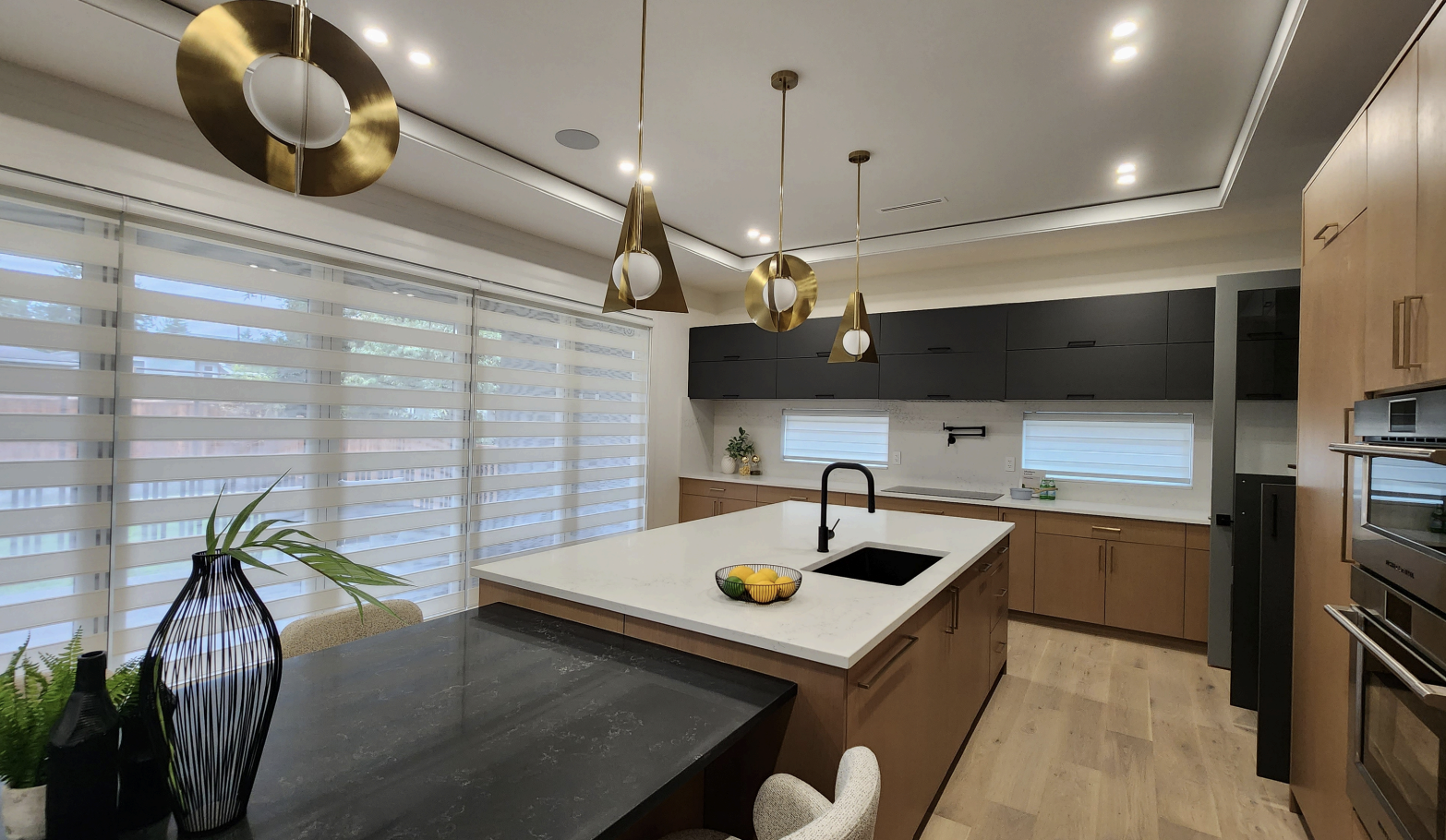 Modern kitchen with a large white island, black sink, black and gold pendant lights, wooden cabinets, black upper cabinets, and large windows with sheer striped curtains.