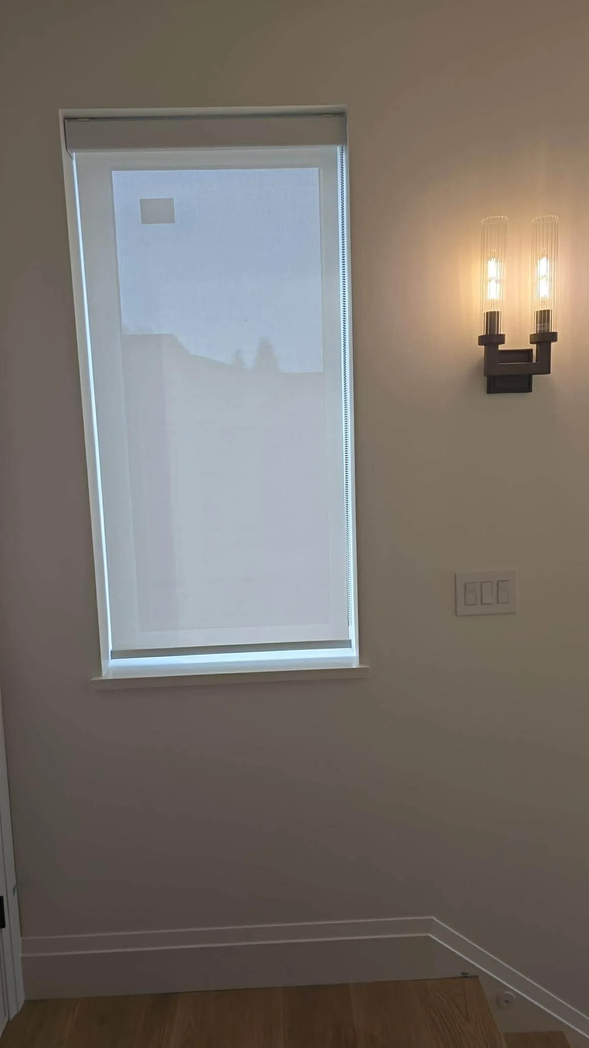 A minimalistic interior wall with a window covered by a roller shade, a wall-mounted modern light fixture with two exposed filament bulbs, and a set of three light switches. The wall is painted a neutral color, and there is wooden flooring at the bottom.