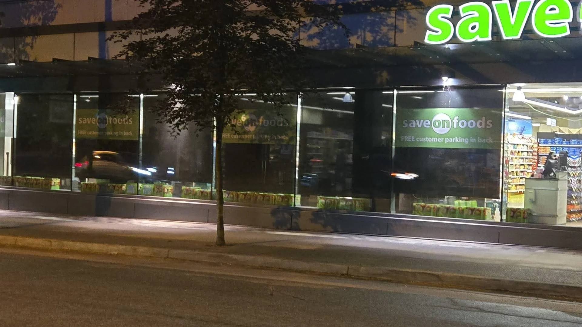 Nighttime exterior view of a grocery store called 'Save On Foods' with signage promoting free customer parking in the back, large windows showing store interior, and a tree in front of the store.